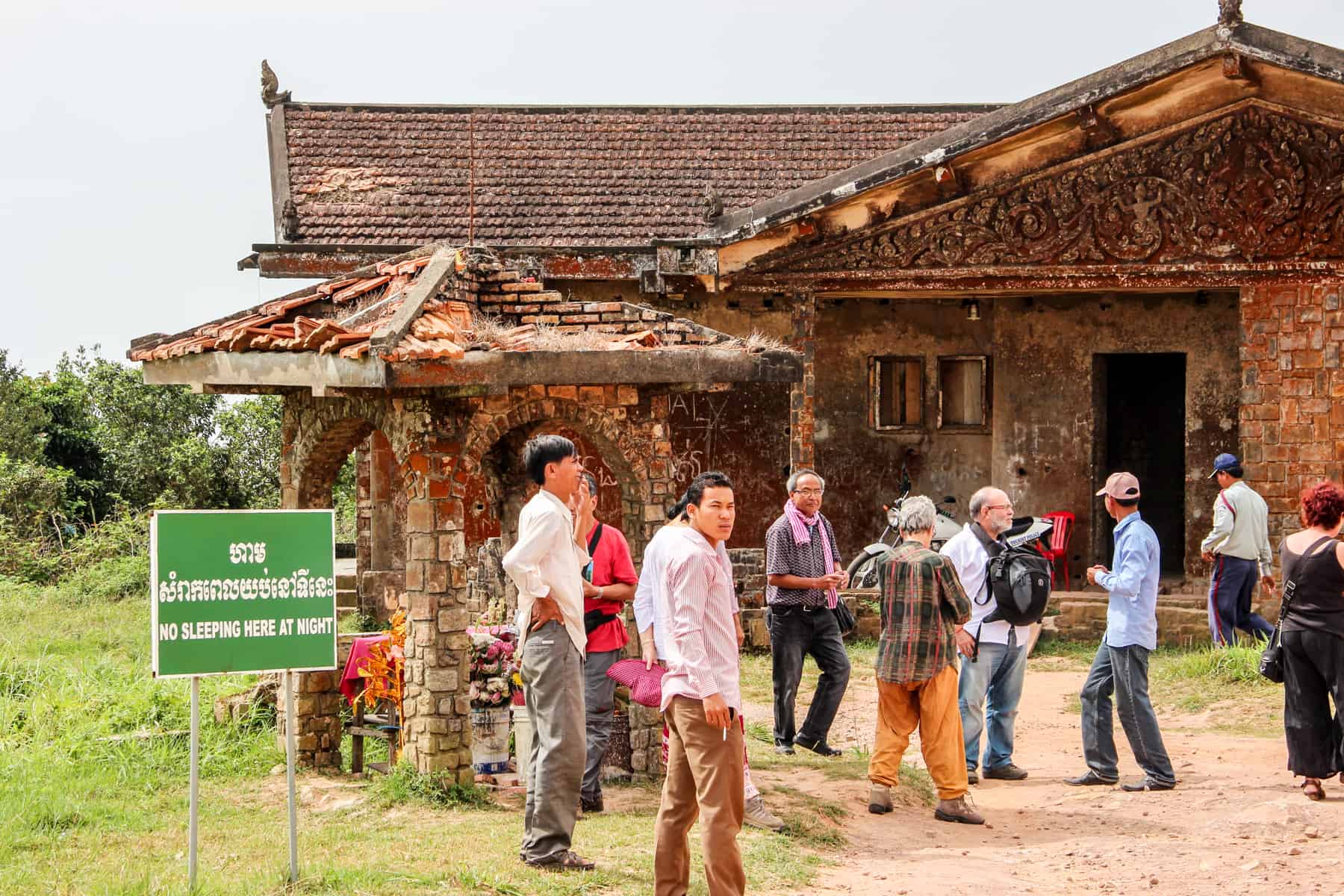A group of people gather outside an abandoned, crumbling, orange brick royal Cambodian palace in Kampot. A green sign says 'no sleeping here at night'.