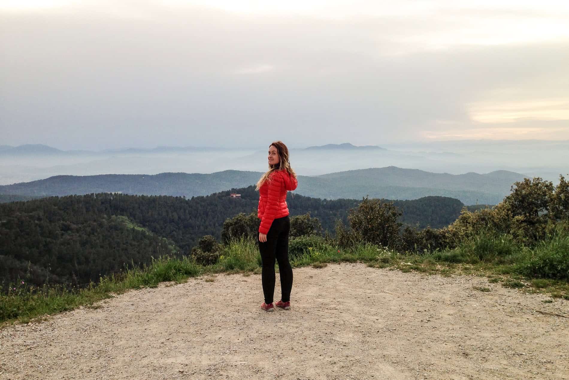 A woman in a red jacket stands on a mountain ledge overlooking the green, coastal, Costa Brava valley.