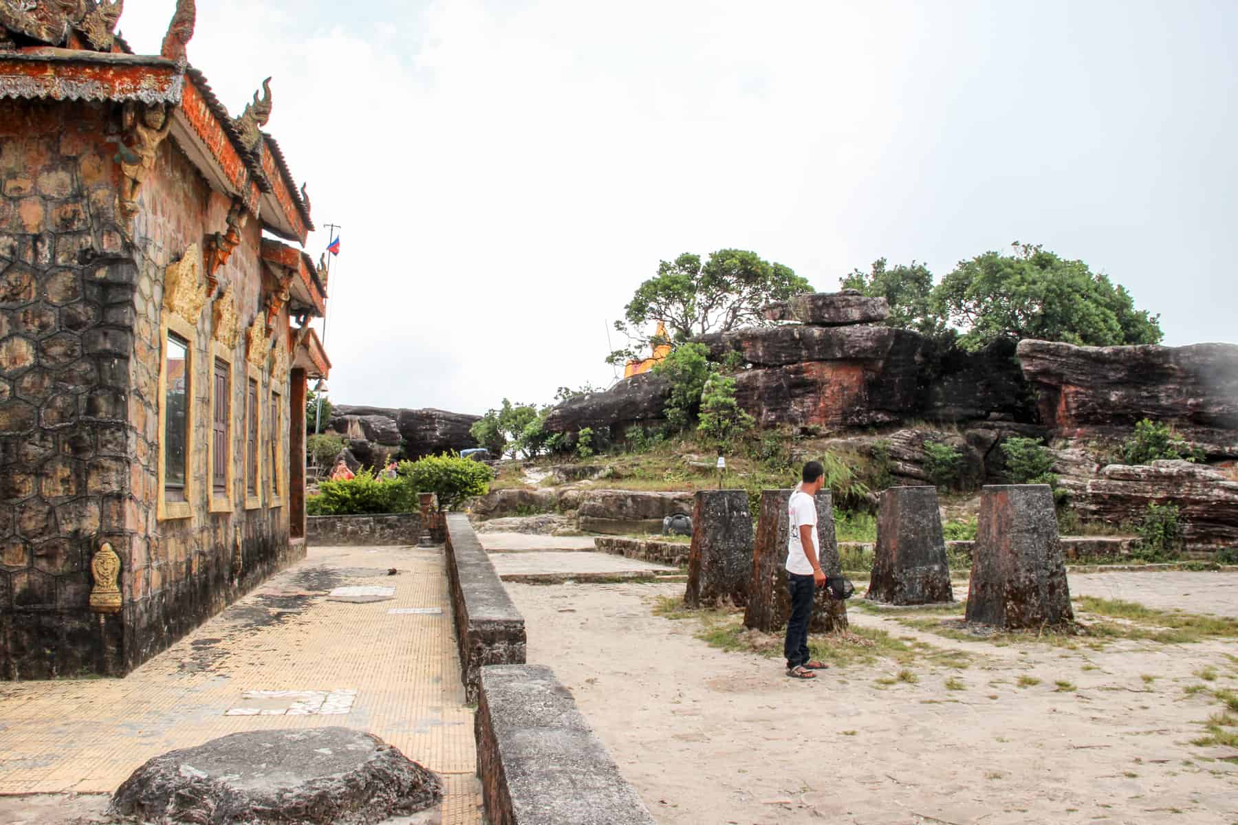 A man in a white t-shirt walks amongst giant black-red stones in front of a run-down, empty stone temple building with yellow and orange trim. 