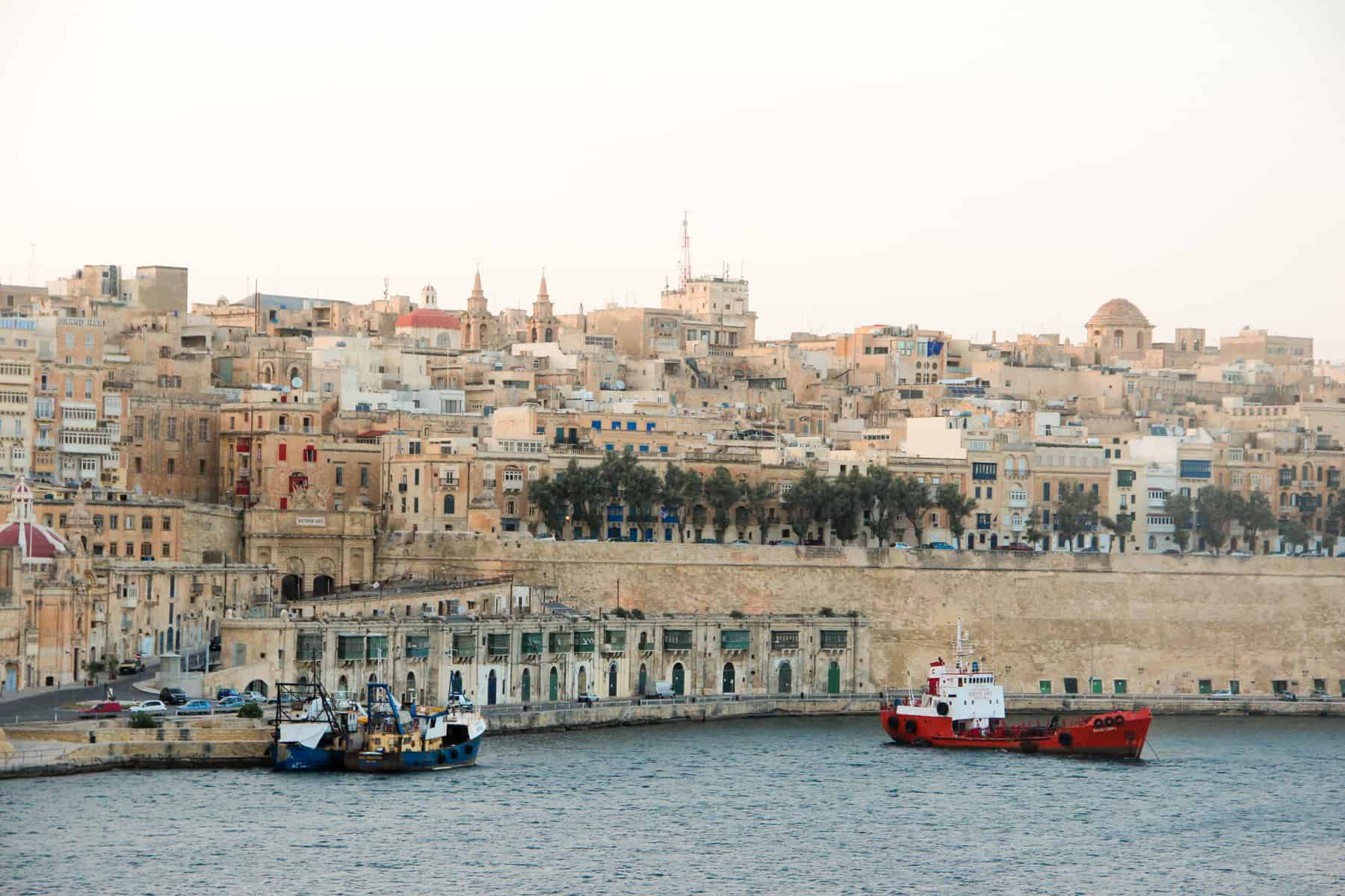 A red and white boat approached a shoreline filled with a wall and rows of old buildings in the same golden caramel hue. 