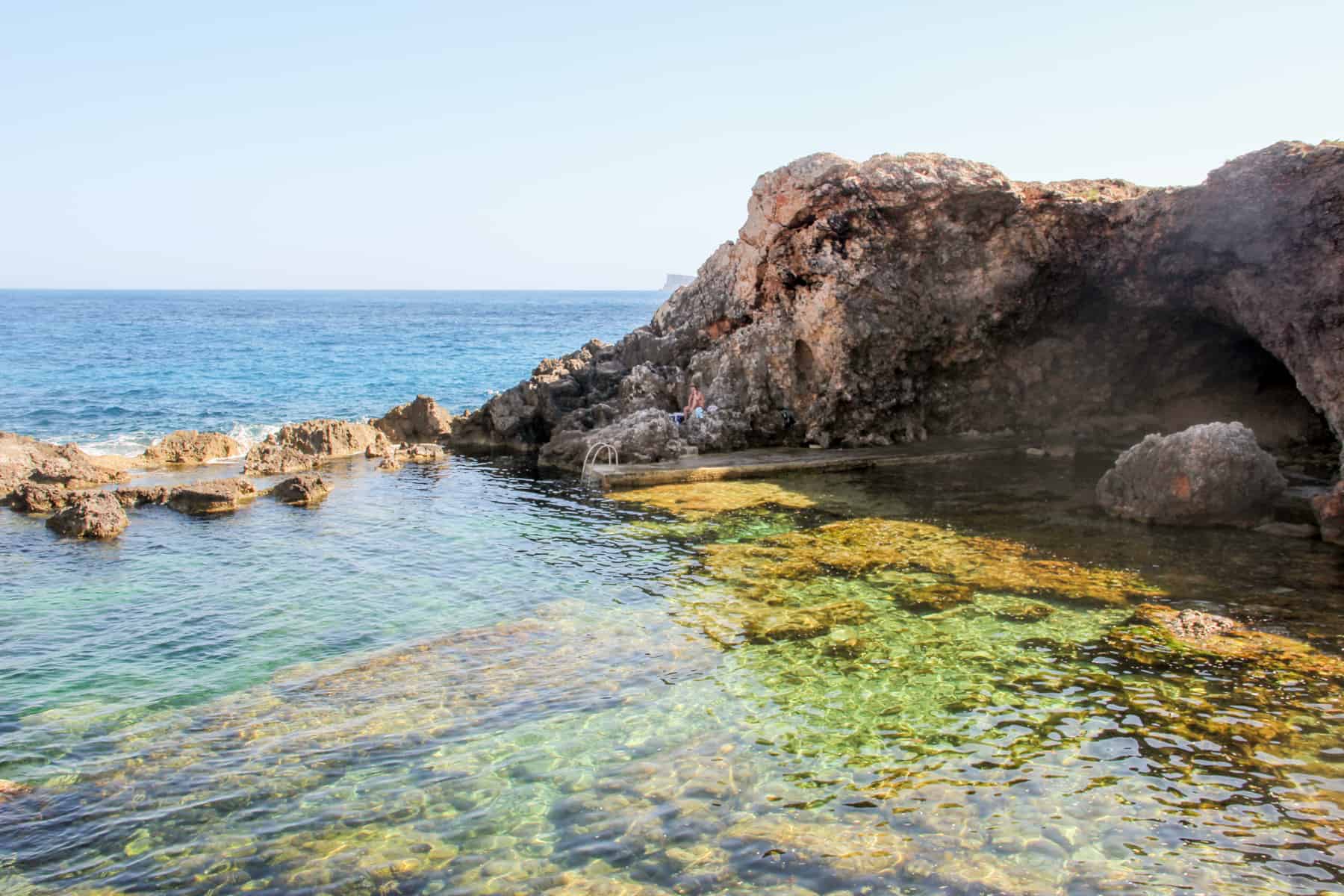 A shallow cove in Malta with blue, green and yellow water, backed by a small curve of rocks. 