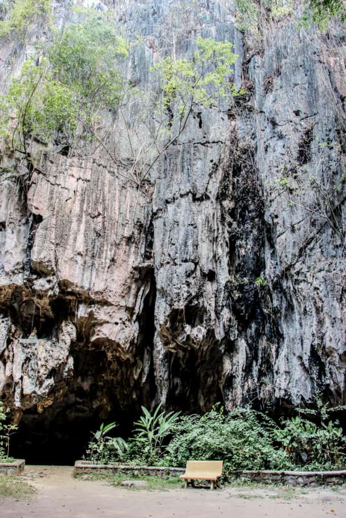 A dark silver high cave wall in with a low dark entrance. A solitary yellow bench sits outside in front of green foliage. 