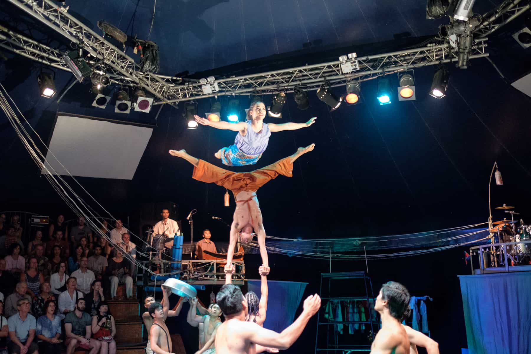 A woman in blue flies over an upside down man doing the splits mid air, held up by a series of performers on the ground. In the background an audience looks on inside the dark circus tent. 