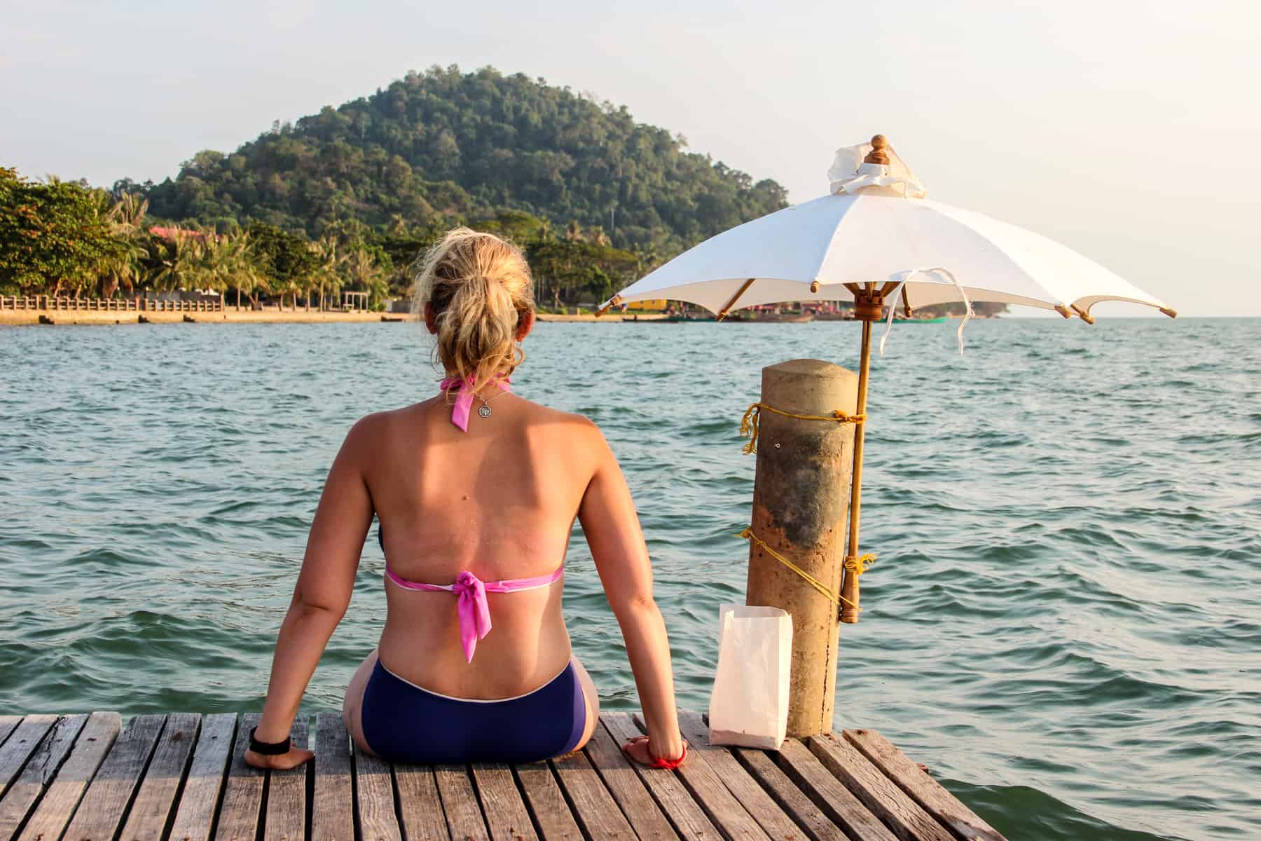 A woman sits on a wooden jetty facing the ocean and the yellow sand coastline of Kep in Cambodia. 