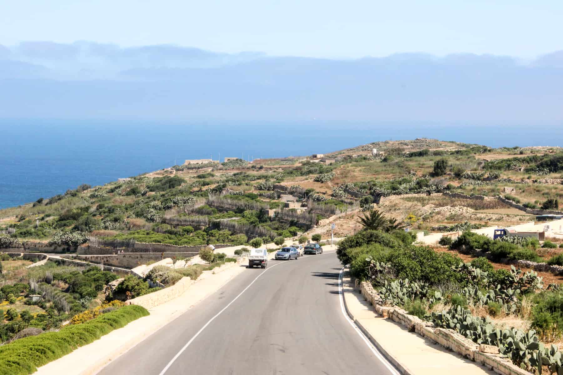 Three cars on a road that bends through the green and golden Malta island landscape, backed by a blue layering of sea, land and sky. 