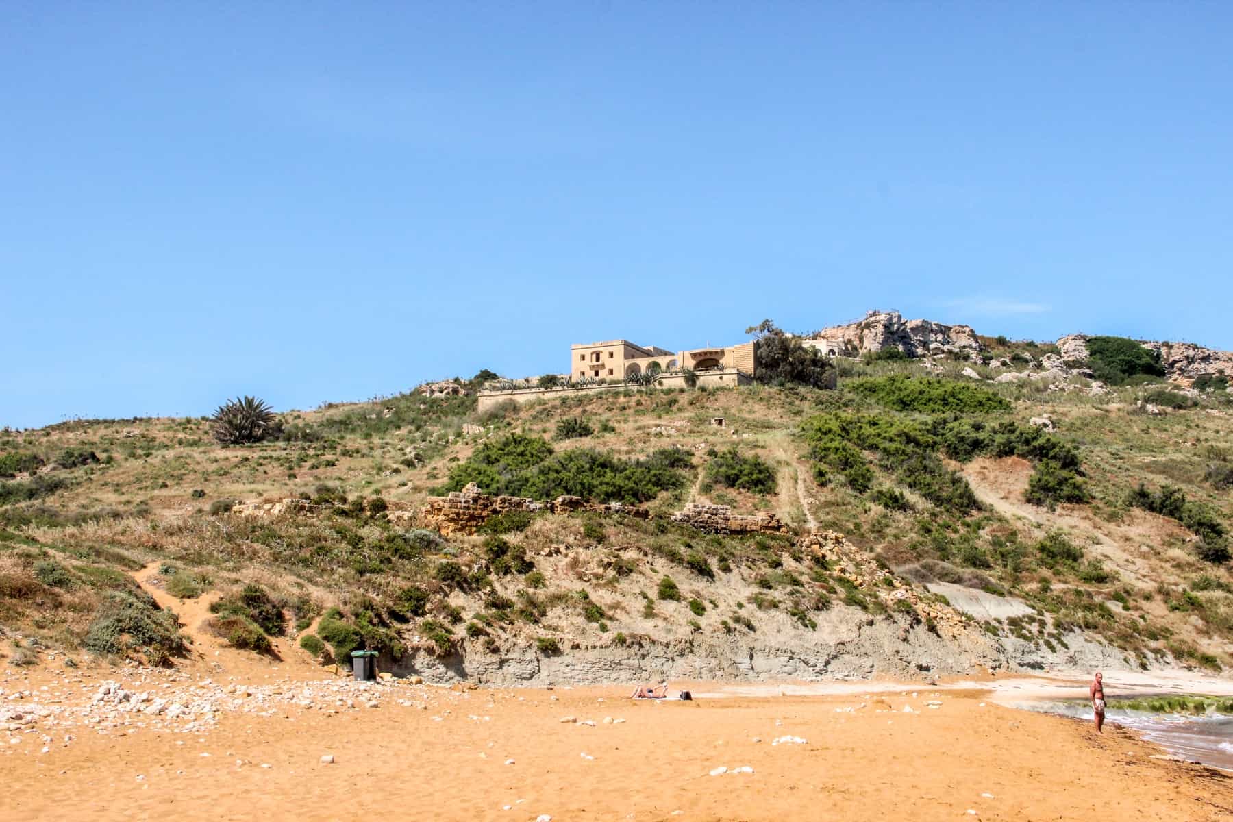 A man stands next to the sea on a golden beach in Malta in front of a verdant hill with a golden house on top. 