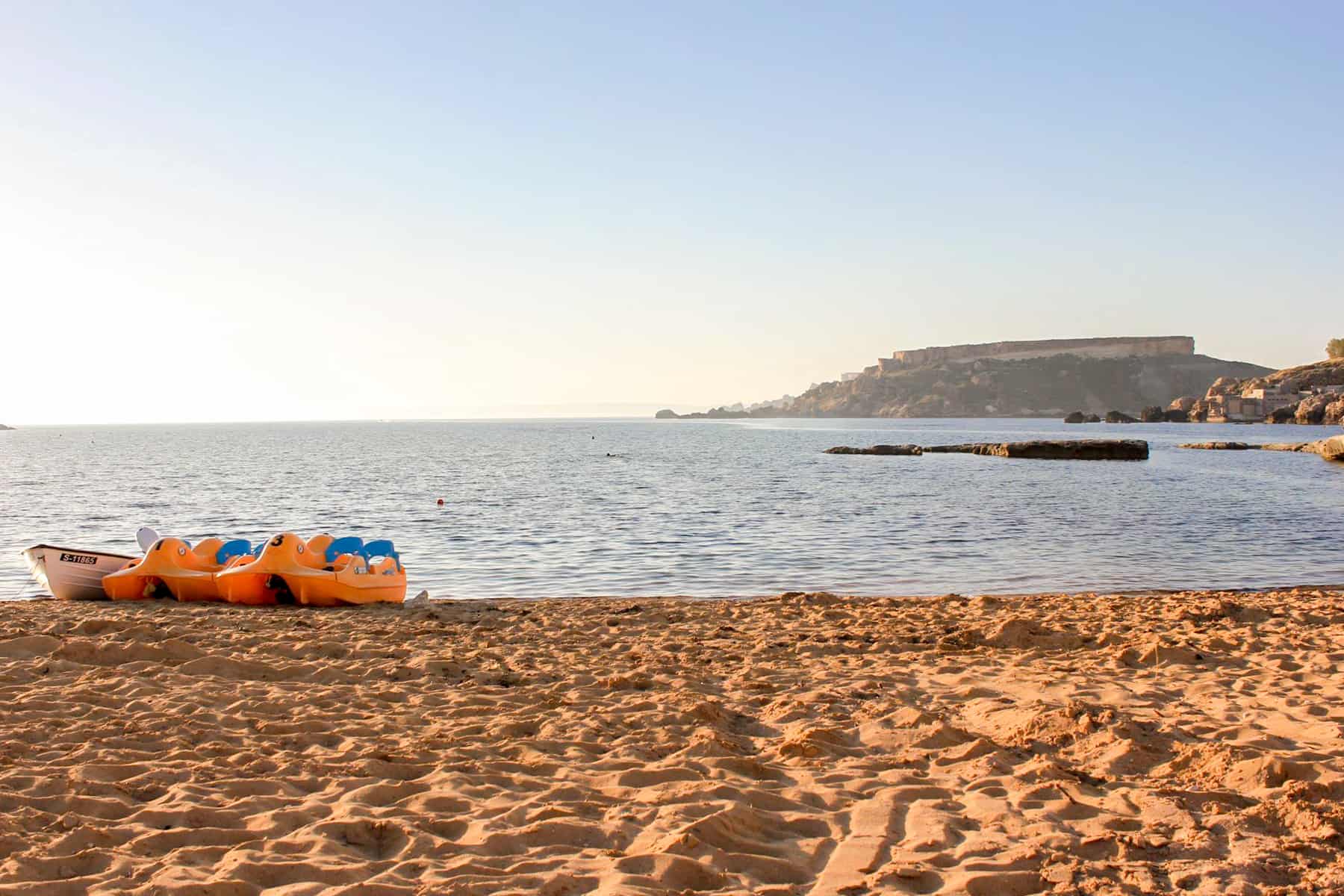 A white boat and two orange peddle boats on a golden sand beach in Malta.