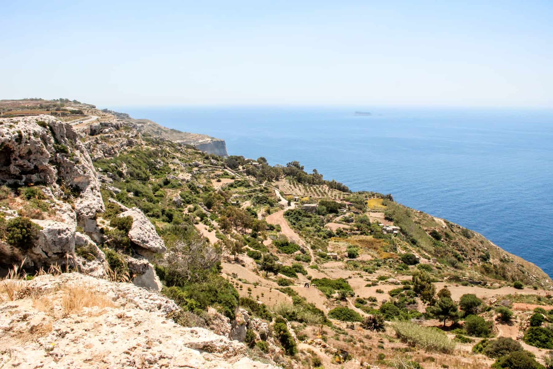 A sloping, rocky cliff face in Malta covered in yellow dusty earth and green shrubs, high above the sea. 