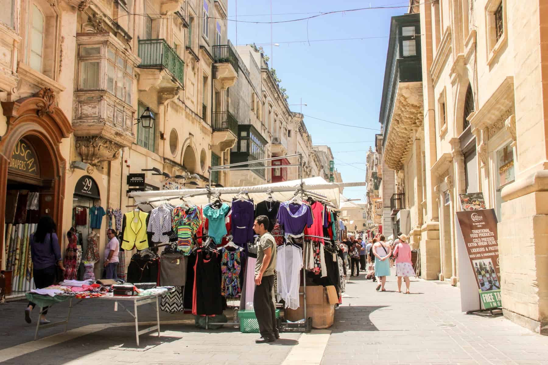 A man stands next to a market stall in the golden streets of Valletta, Malta, selling clothing in various bright colours. 