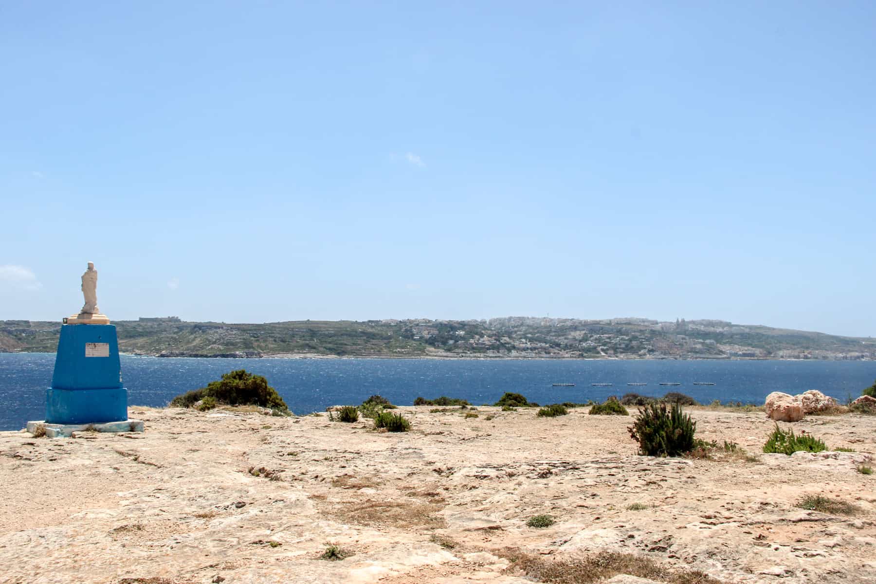 A small white statue of Jesus on a blue stand - known as the little chapel of Malta - perched on a rocky cliff face, overlooking the sea.