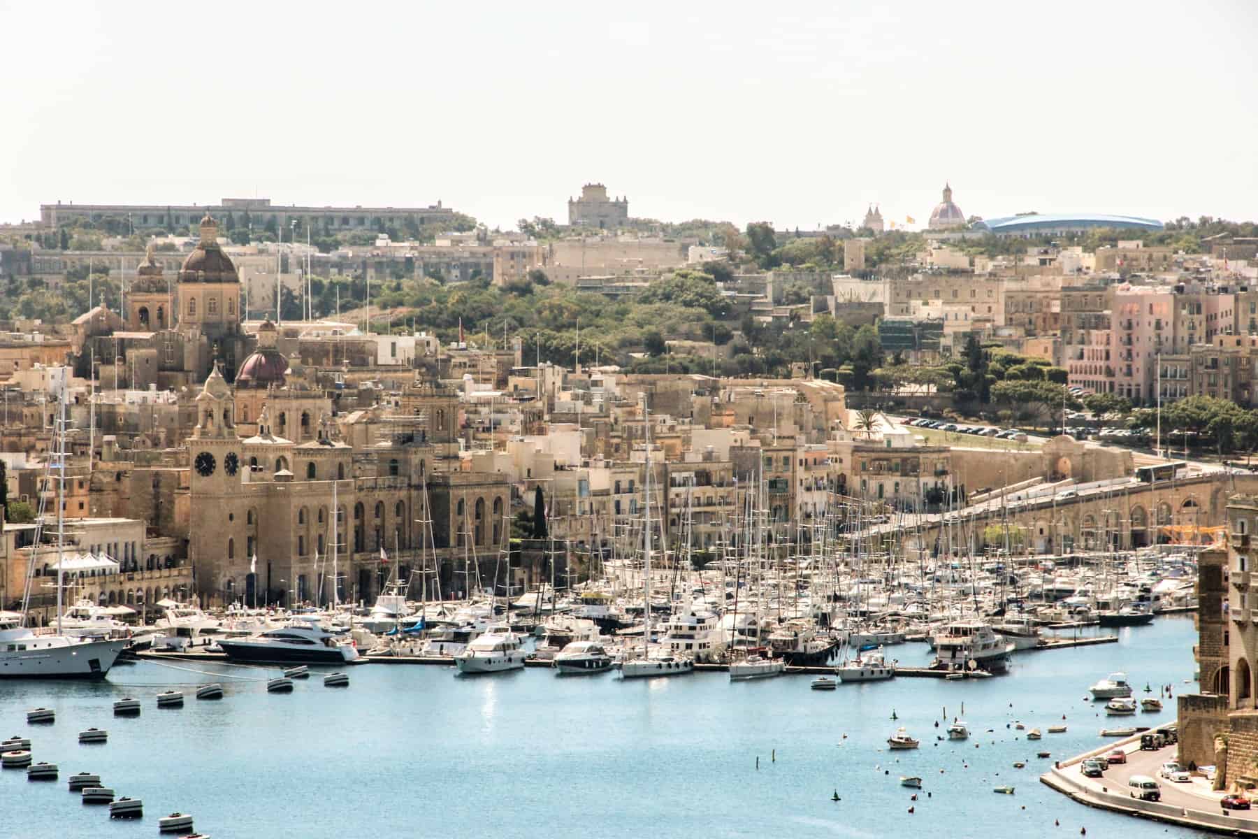 A sky blue water sea port in Malta full of ships and backed by old, golden stone buildings reaching all the way to the hilltop.