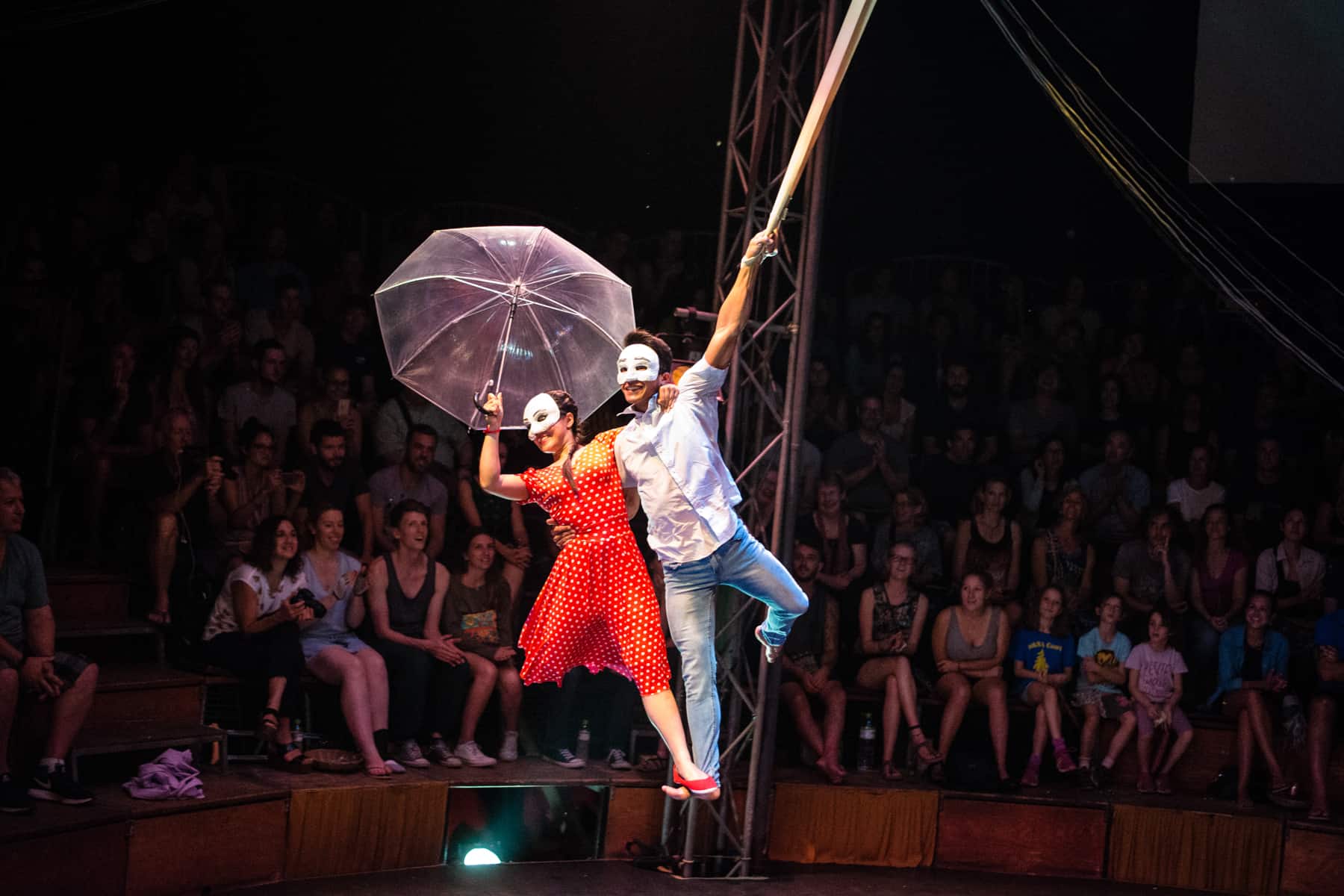 A white-masked woman in a red dress, holding an umbrella and a white-masked man man in blue jeans an a white top holding onto a rope perform a mid-air acrobatic performance in front of a audience in a circus in Siem Reap.