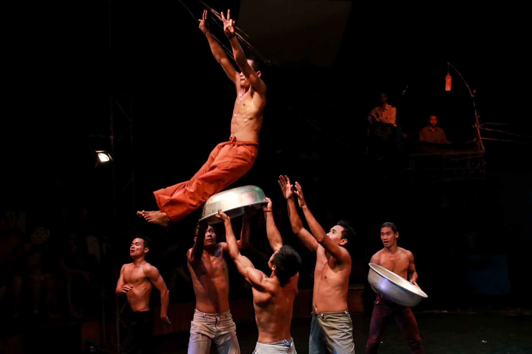 Three men hoist another into the air using a metal bowl during a circus performance in Cambodia. Two other men can be seen in the background, another holding a large silver bowl.