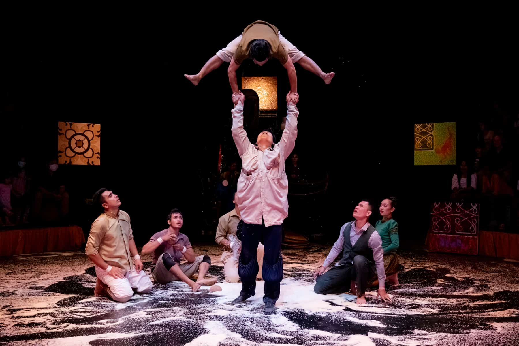 Five performers sit on the ground covered in rice grains looking up at a man balancing another high above his head. The background is dark with golden lighting.