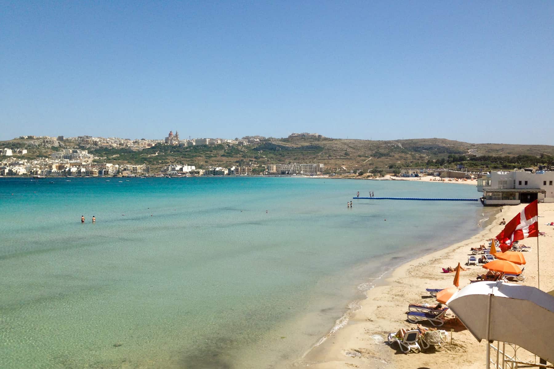 The Maltese flag(red with a white cross) flies over a long strip of yellow sand beach that curves around the coastline towards the green, building-filled hills. on a wide coastline 