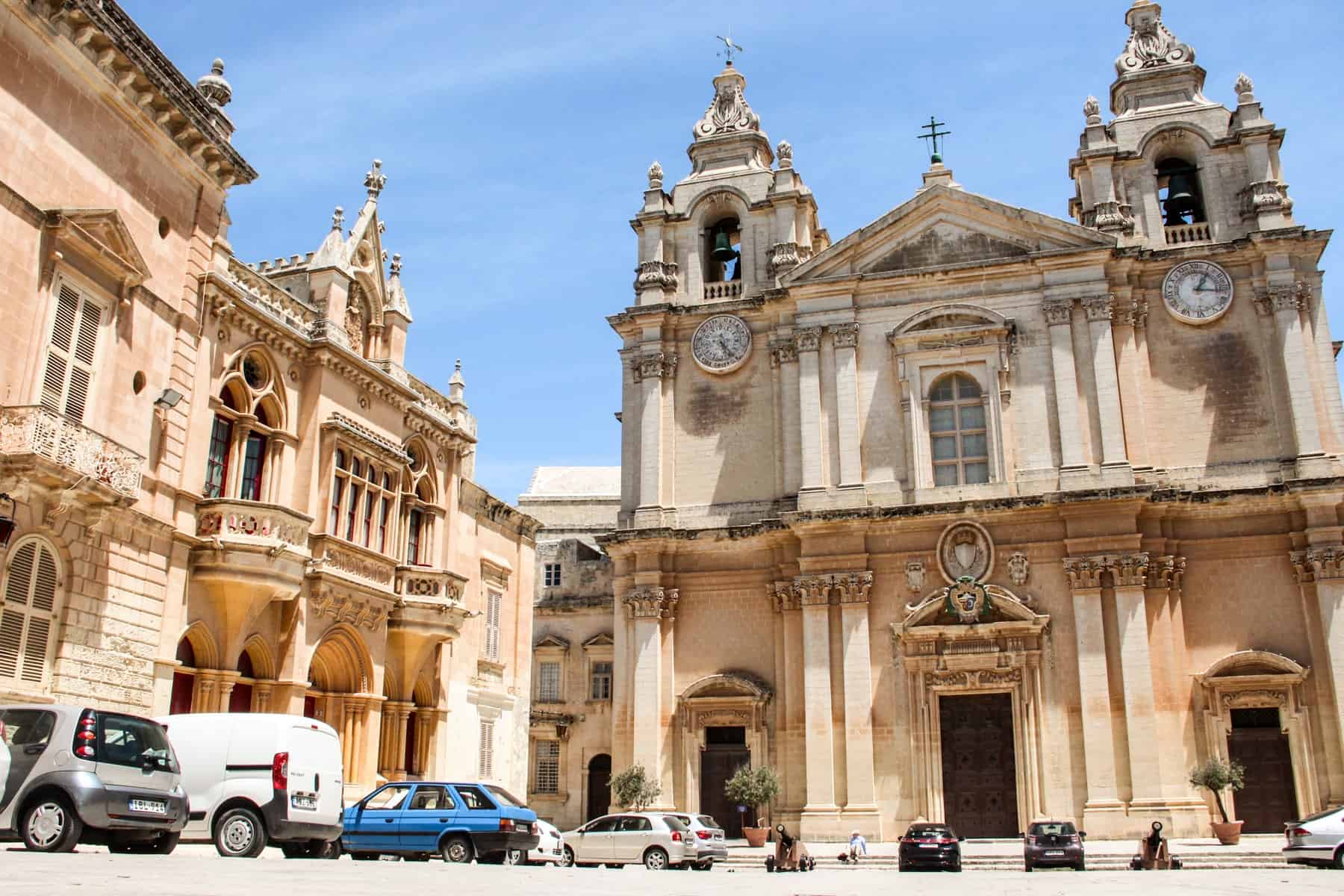Grey, white and blue cars outside a corner of majestic golden buildings with columns, towers and spires in Mdina, Malta.
