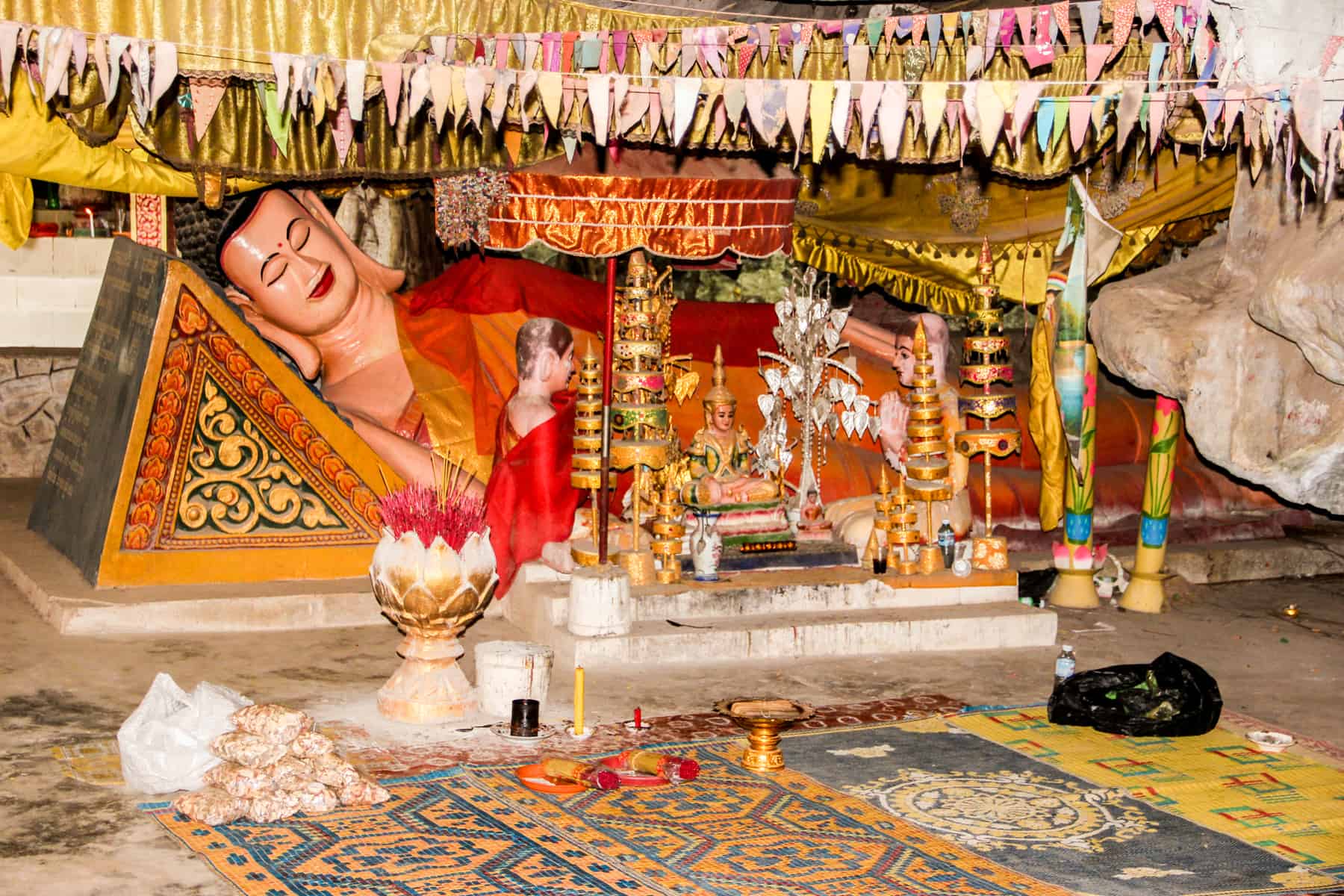 A reclining Buddah statue inside a cave, surrounded by rugs and prayer offerings.