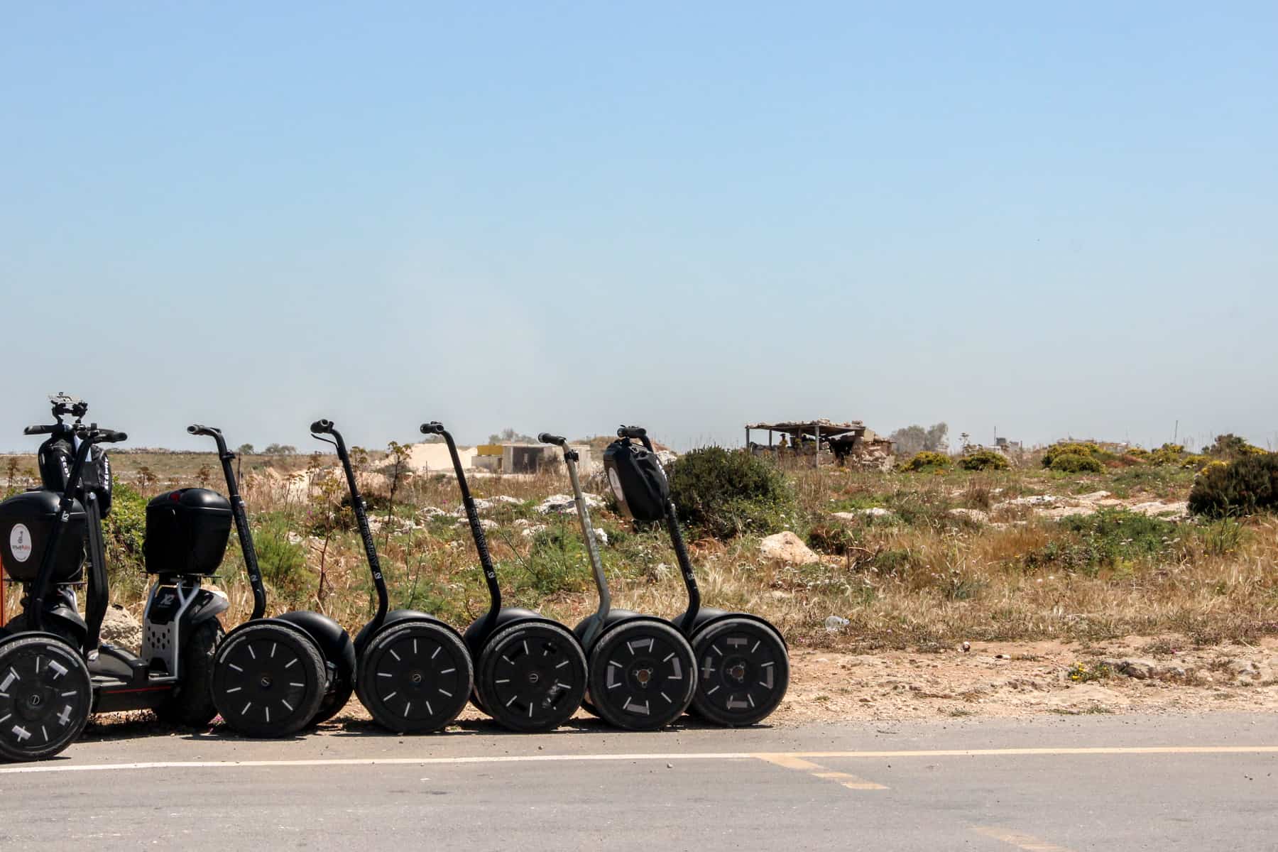 Six black segways lined up on a road next to messy grassland, ready for a trip in Malta. 