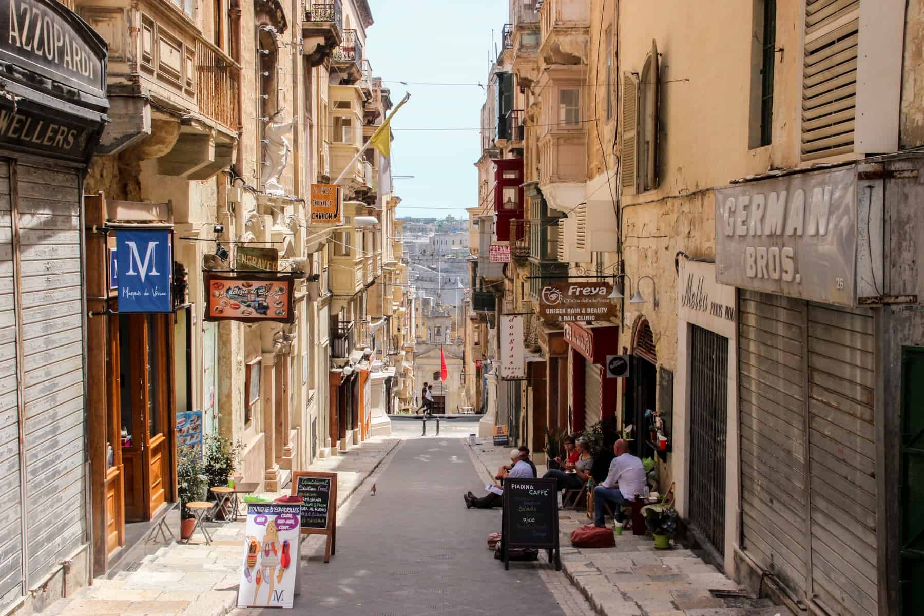 Five people sit outside a cafe on the pavement in a narrow, golden, shop-sign filled street in old Valletta in Malta island. 