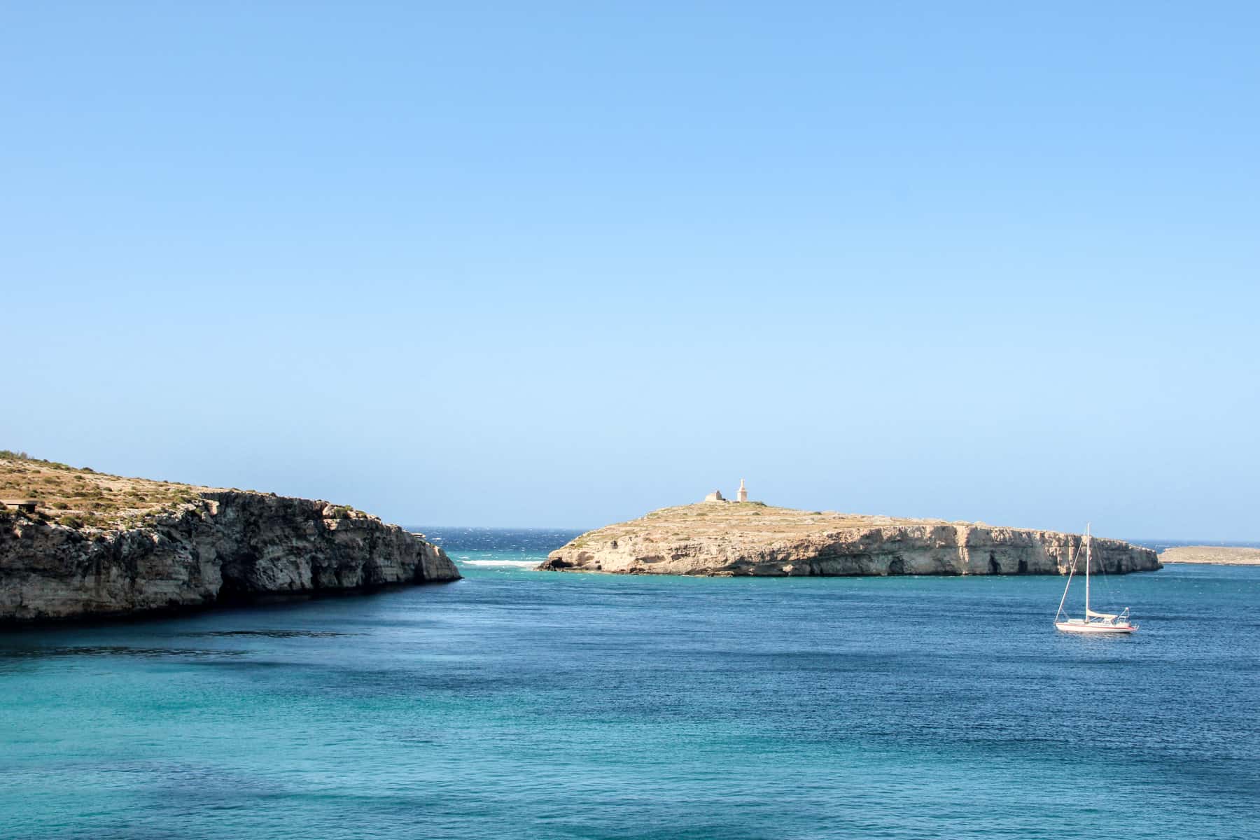 A small, white sail boat on the two-tone blue waters in Malta, heading towards a gap between two low-lying rocks. 
