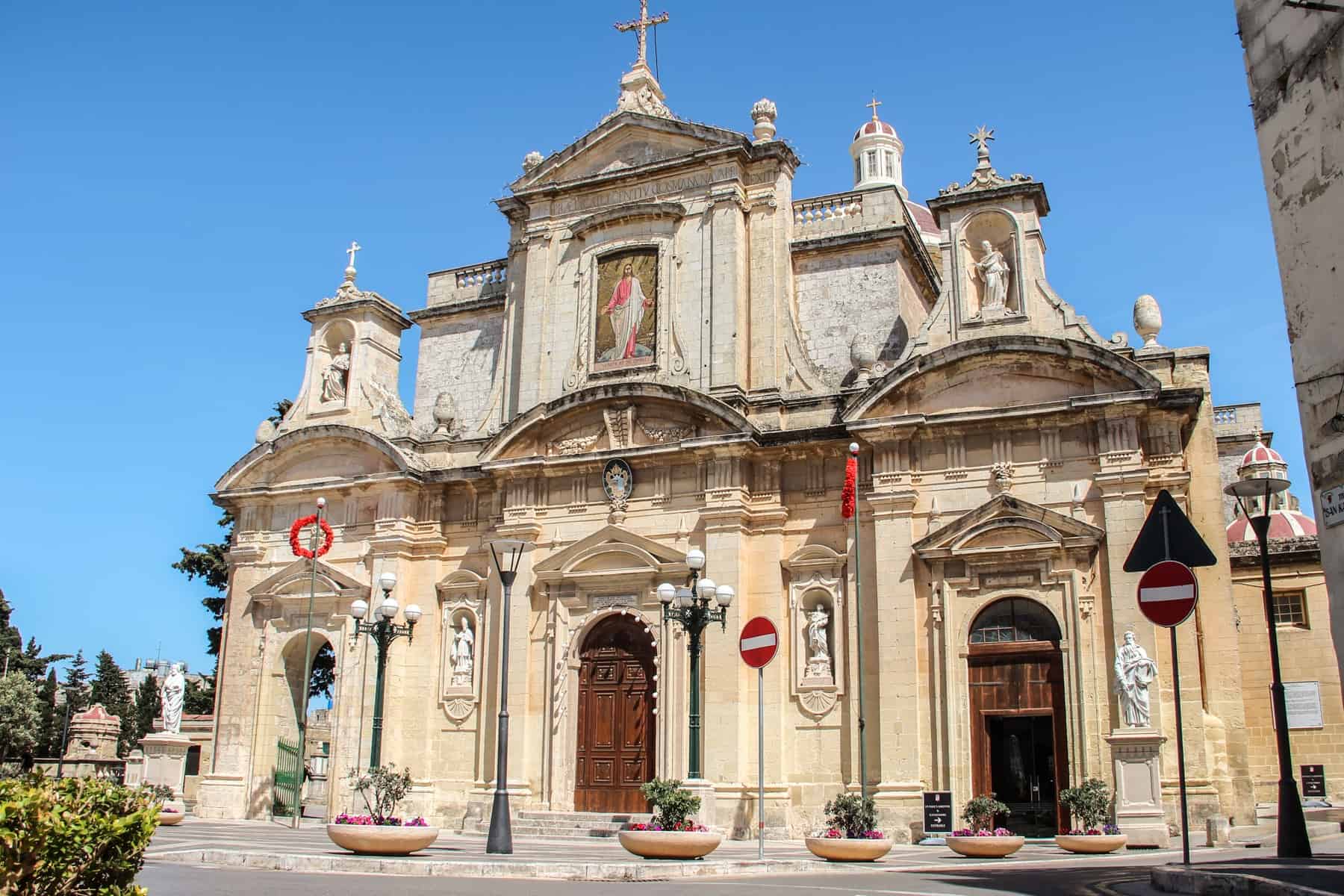 A yellow stone church in Malta with three arched doorways and a rooftop turret. 