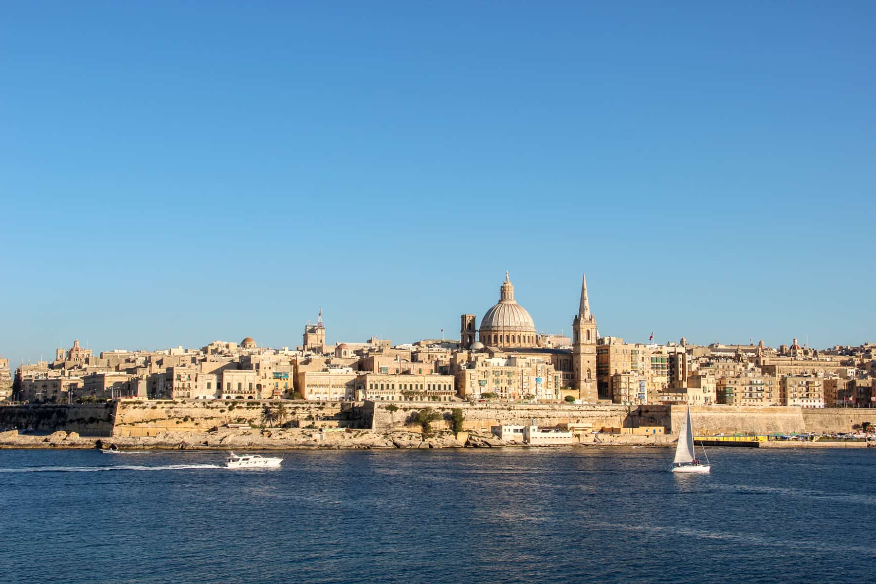 A view to the golden structures, spires and domes of Valletta, Malta. The far view is taken from the sea, where a white boat and a white sailing boat pass by.