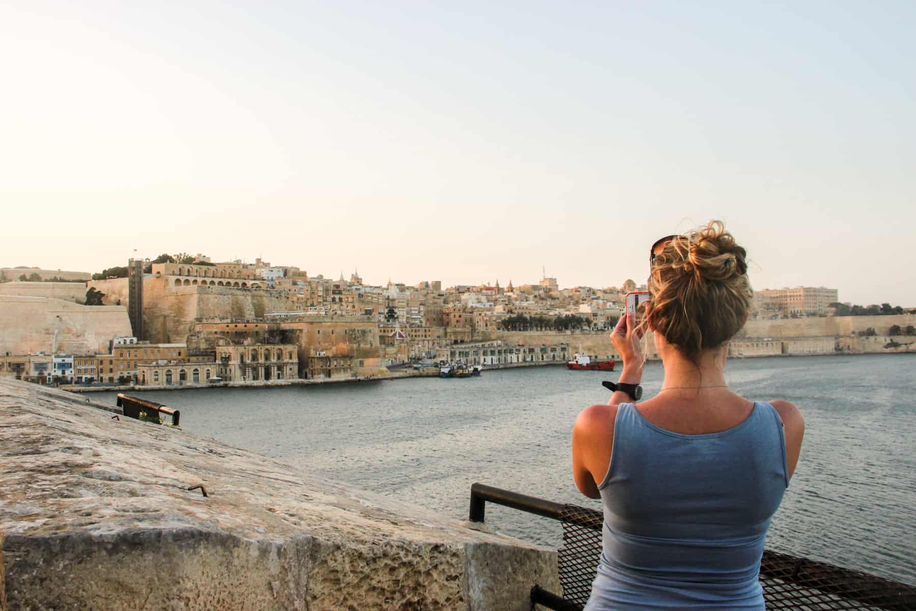 A woman in a blue vest top takes a picture with her mobile phone of the golden architecture view to Valletta in Malta. 
