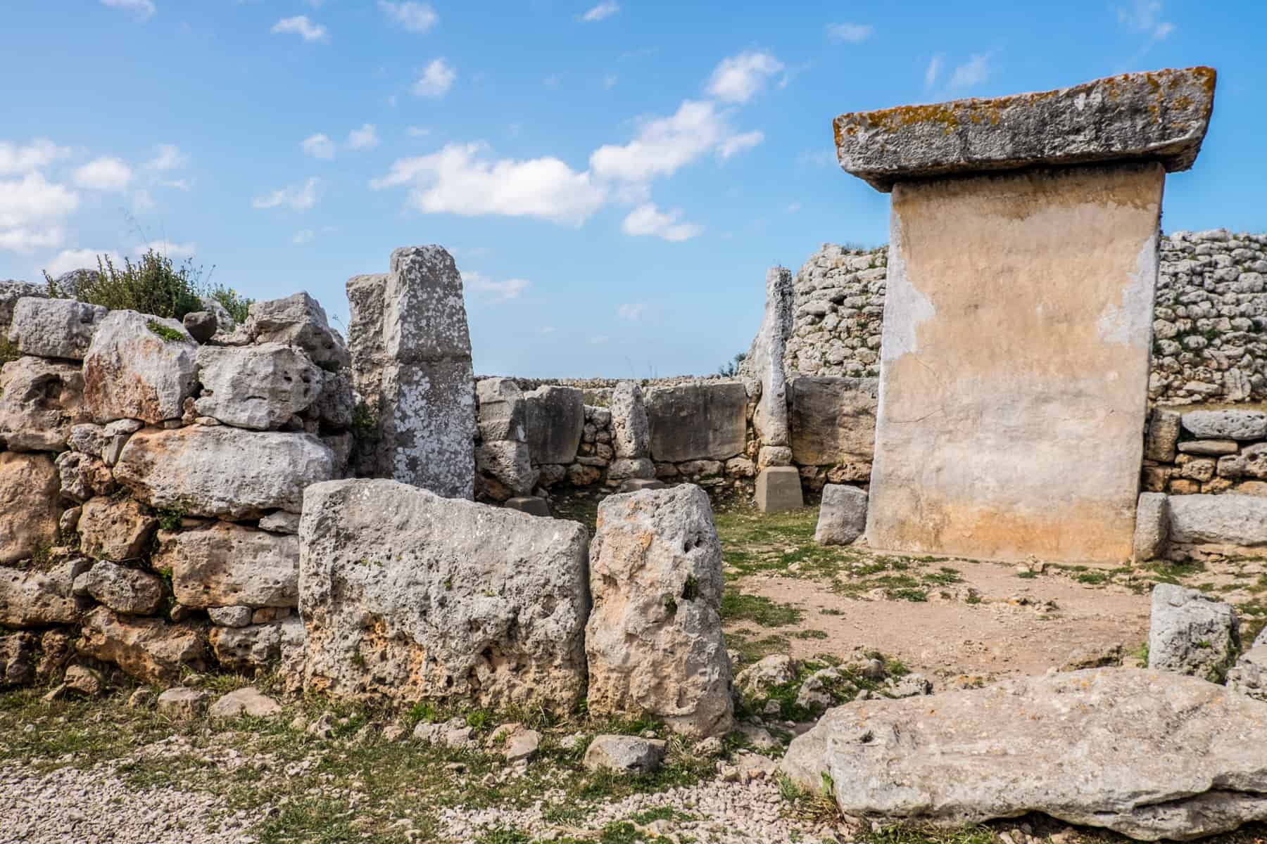 A tall glden rock slab with a flat, silver rock on top stands within an area surrounded by other ancient rocks - one of the Megalithic monuments on the island of Menorca. 