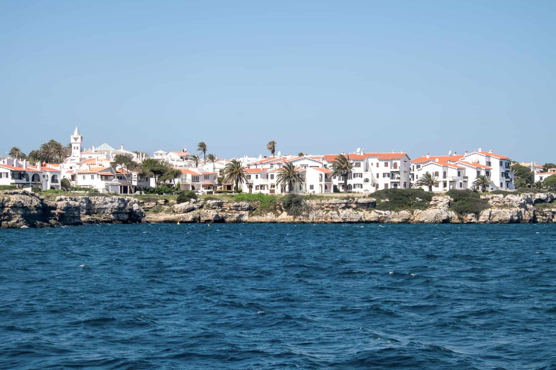 View from a boat on dark blue waters looking towards a rocky coastline filled with luxury white houses with orange rooftops. 