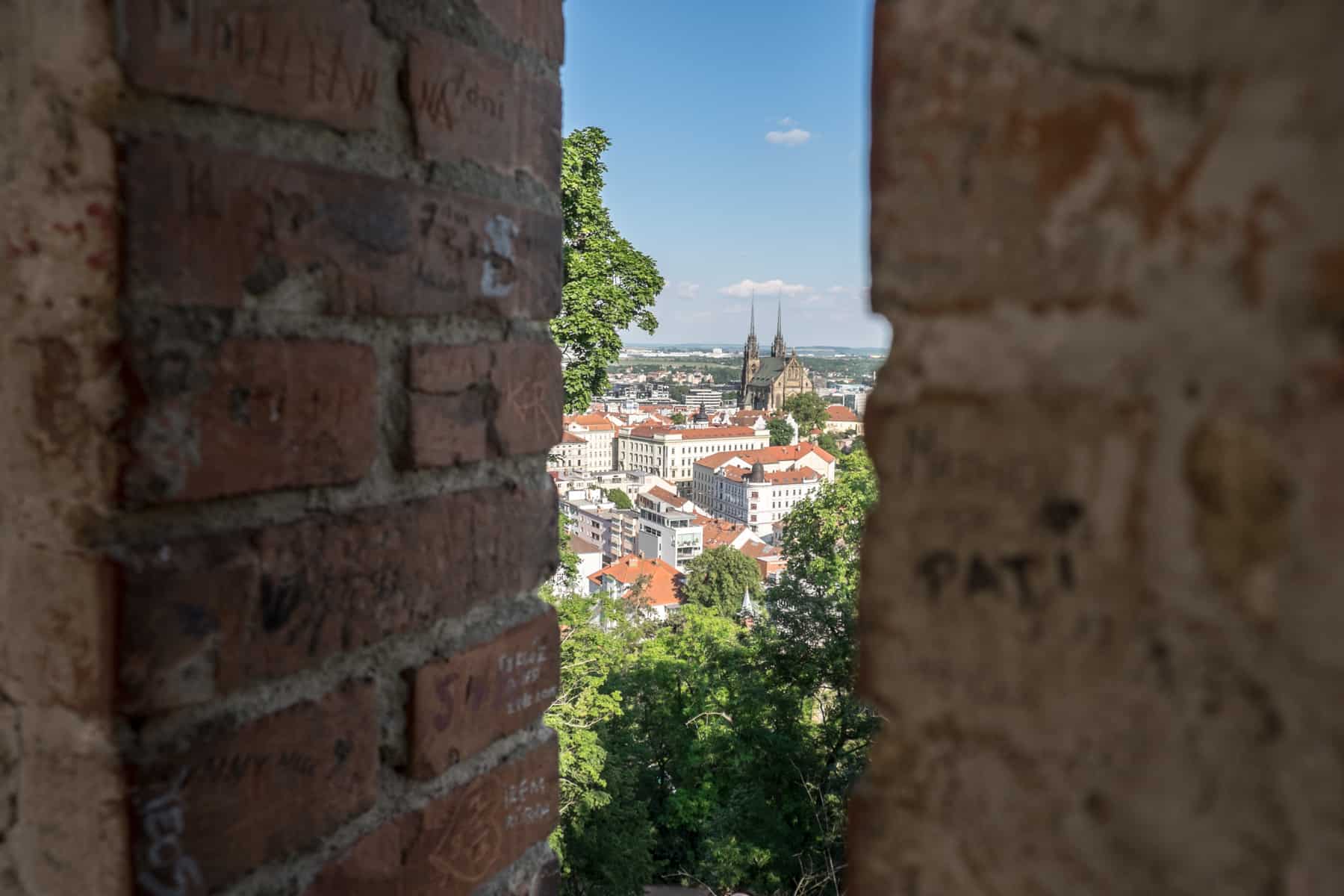 A view to Brno's caramel-black Cathedral, and the surrounding red roofed city buildings, from between the brick walls of a castle tower at a higher level.