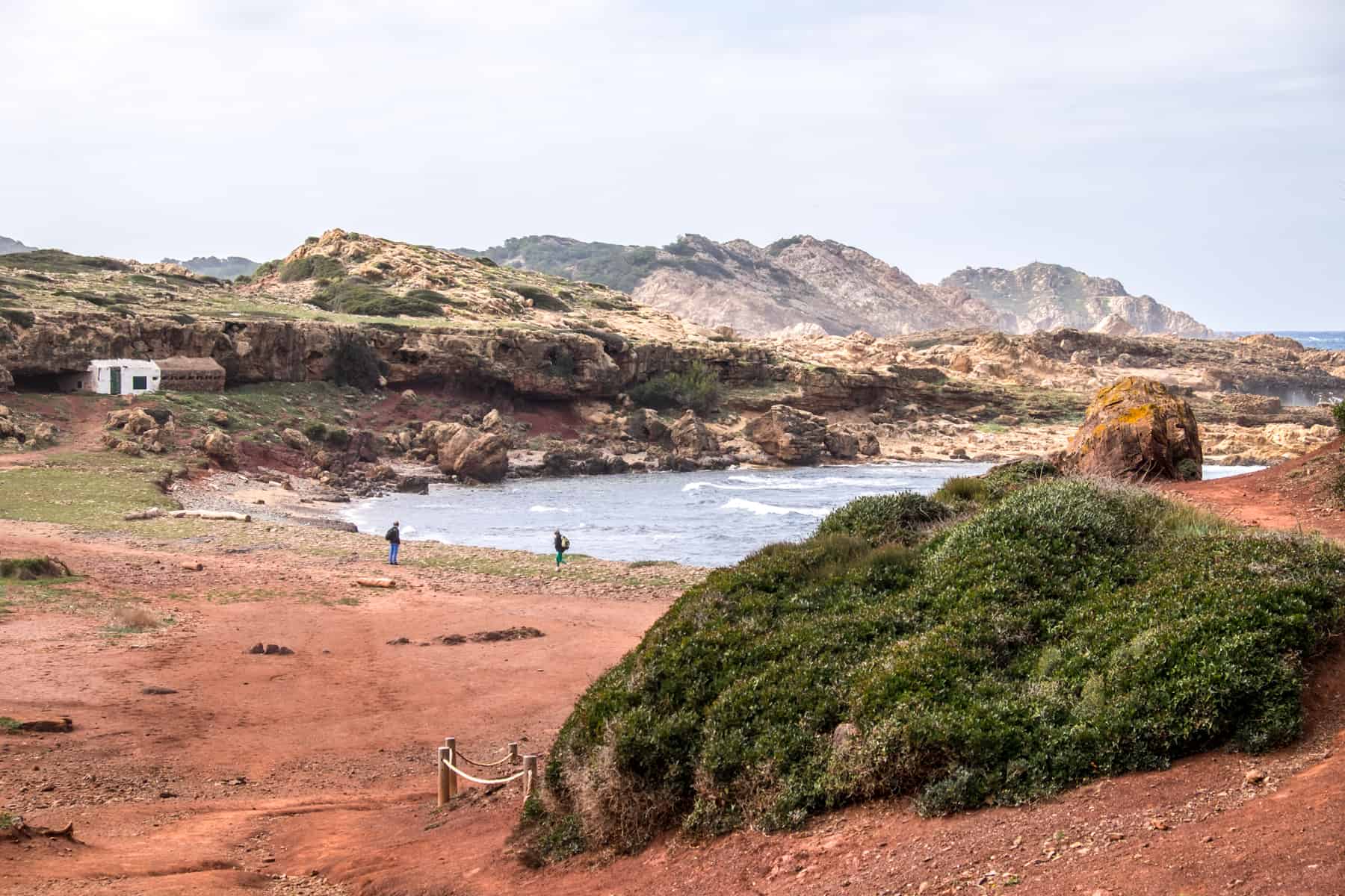 An ochre red beach cove with a small bay of water, surrounded by jagged rocks covered in green. 