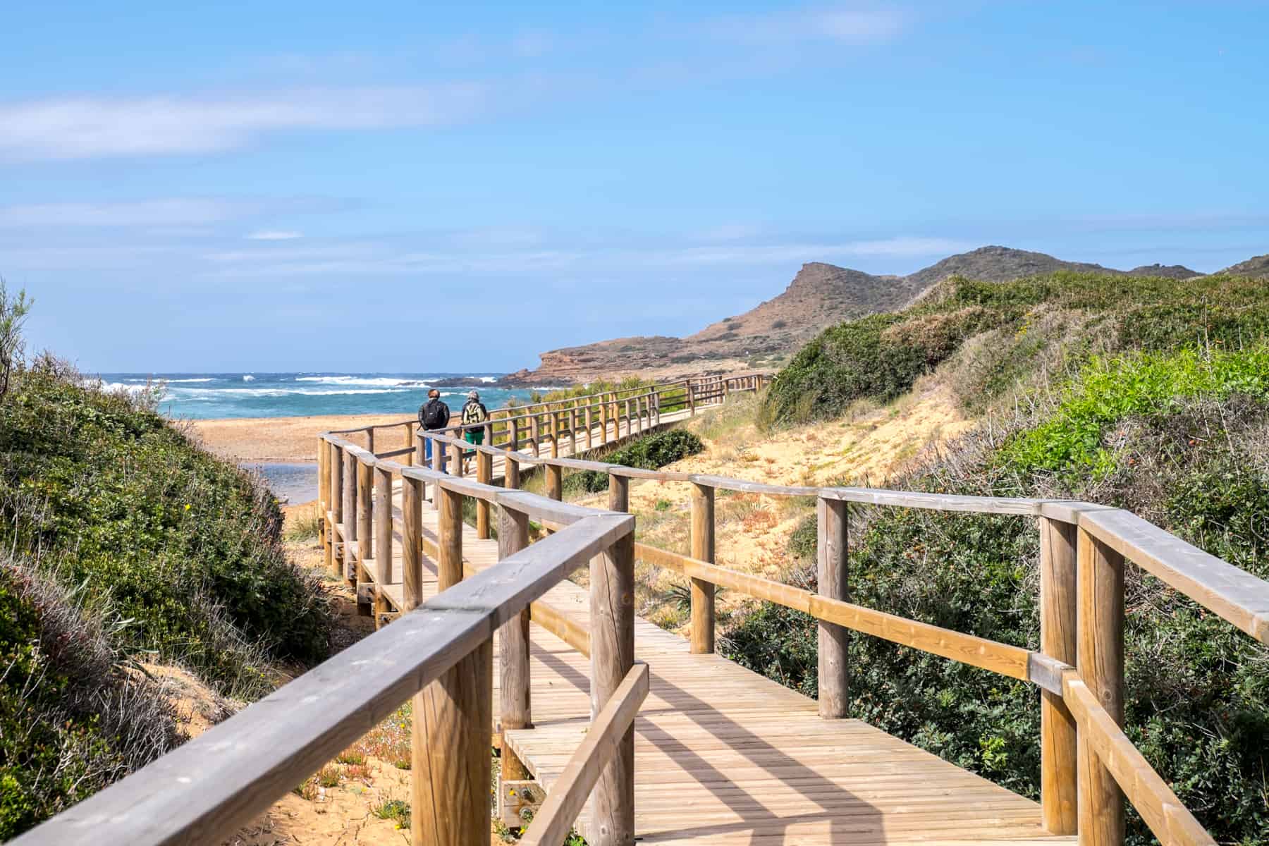 Two people walking on an elevated wooden walkway over a yellow sand beach, backed by grassy hills.