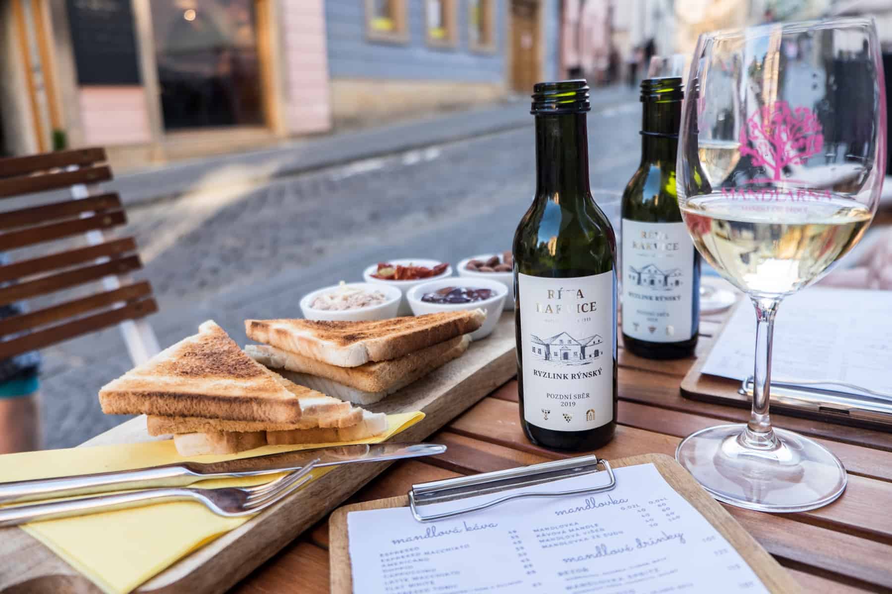 Toast and snack, two small bottles of Moravian wine and a glass of white wine on a wooden table next to a pastel building street. 