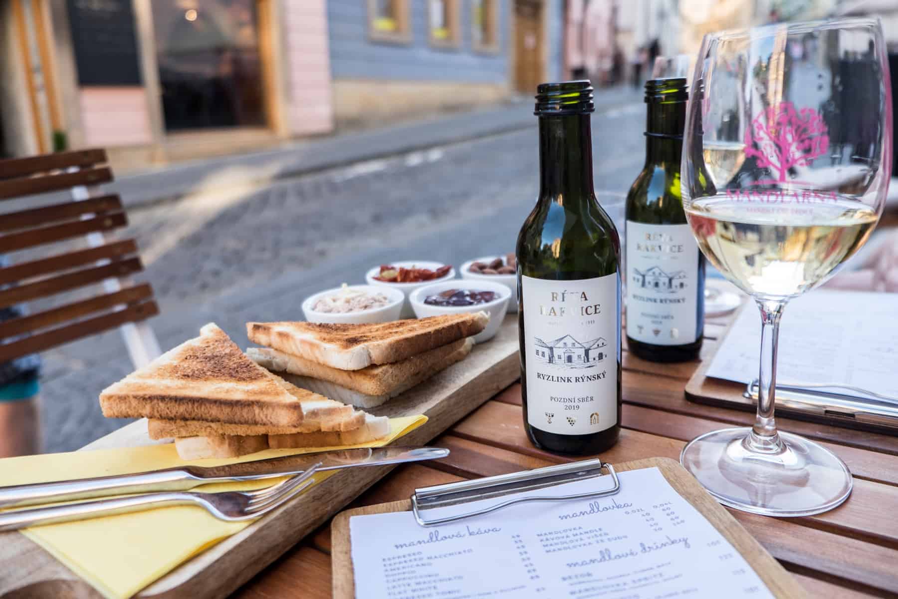 brno places to visit Toast and snack, two small bottles of Moravian wine and a glass of white wine on a wooden table next to a pastel building street.