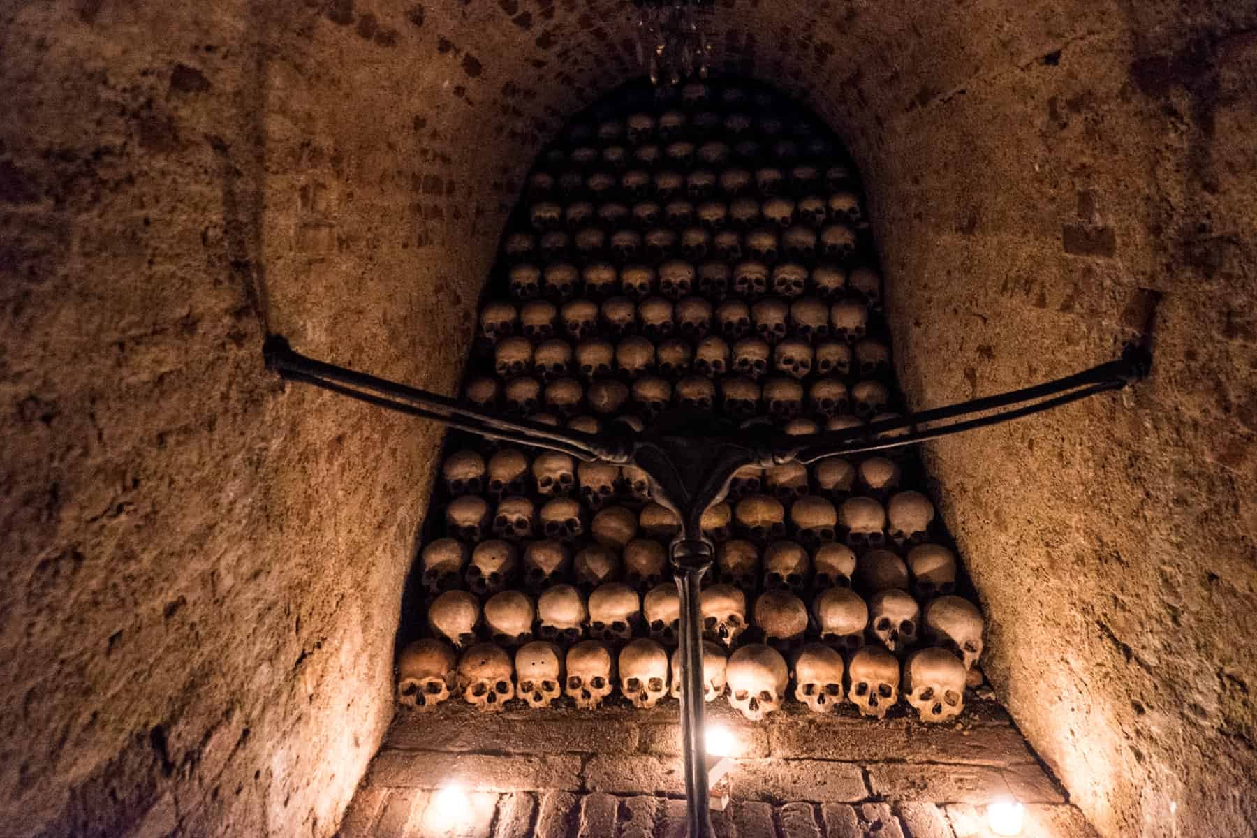 At the Ossuary at the Church of St James in Brno, rows of skulls are pilled line by line on top of one another at the end of a tunnel, illuminated by golden light. 