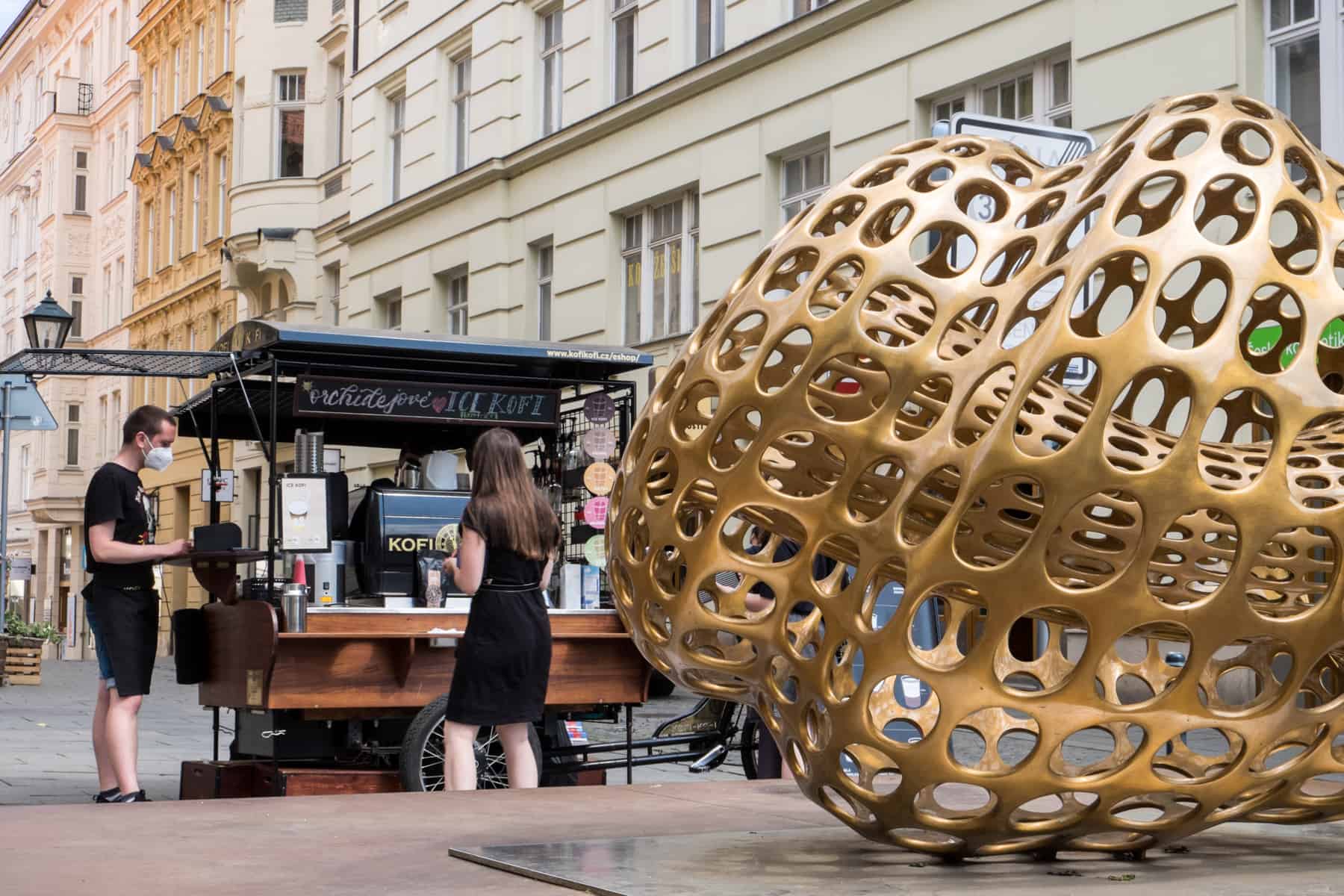 A man and woman stand at a black and dark wood mobile coffee cart that stands behind a giant golden art sculpture in Brno.