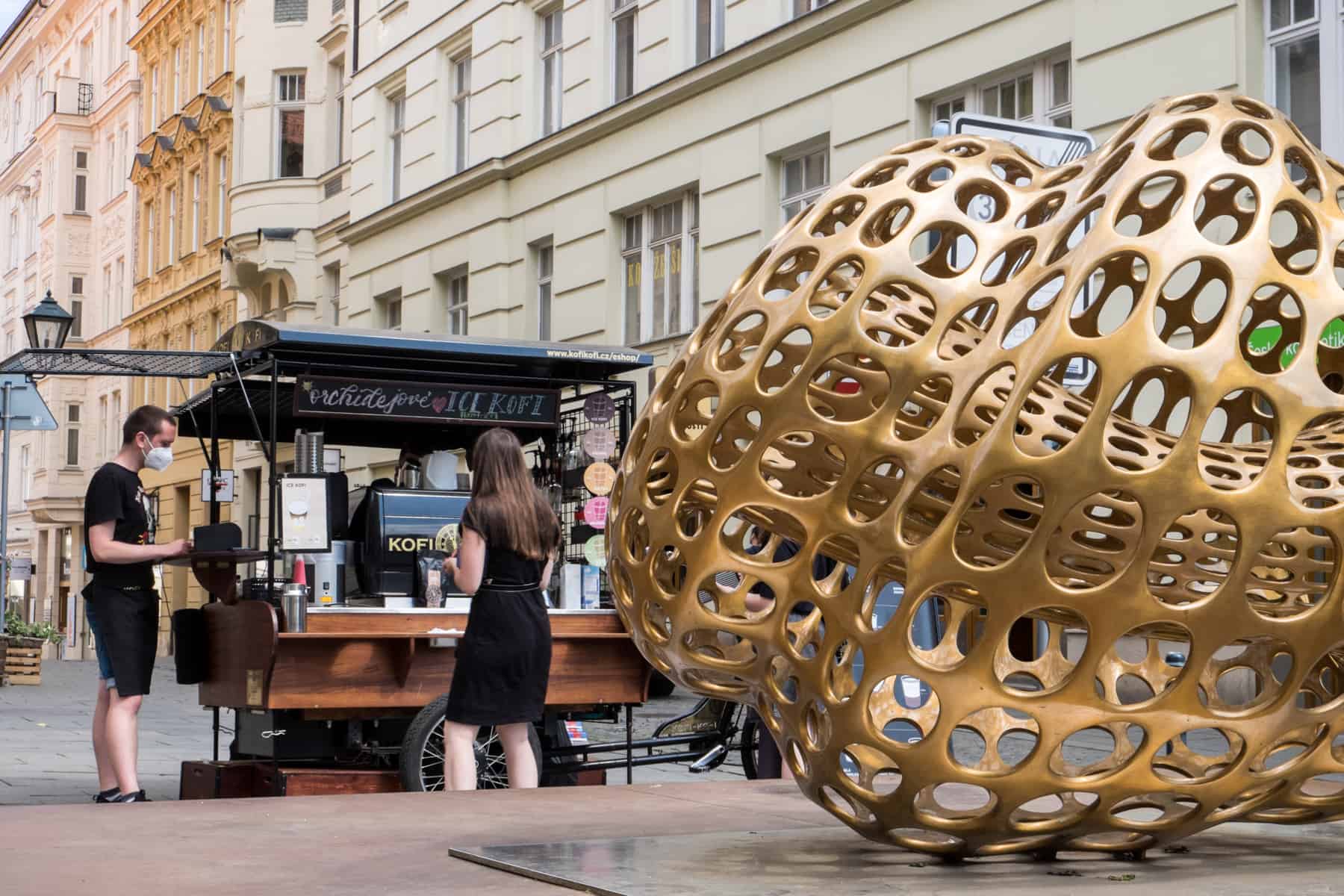 brno places to visit A man and woman stand at a black and dark wood mobile coffee cart that stands behind a giant golden art sculpture in Brno.