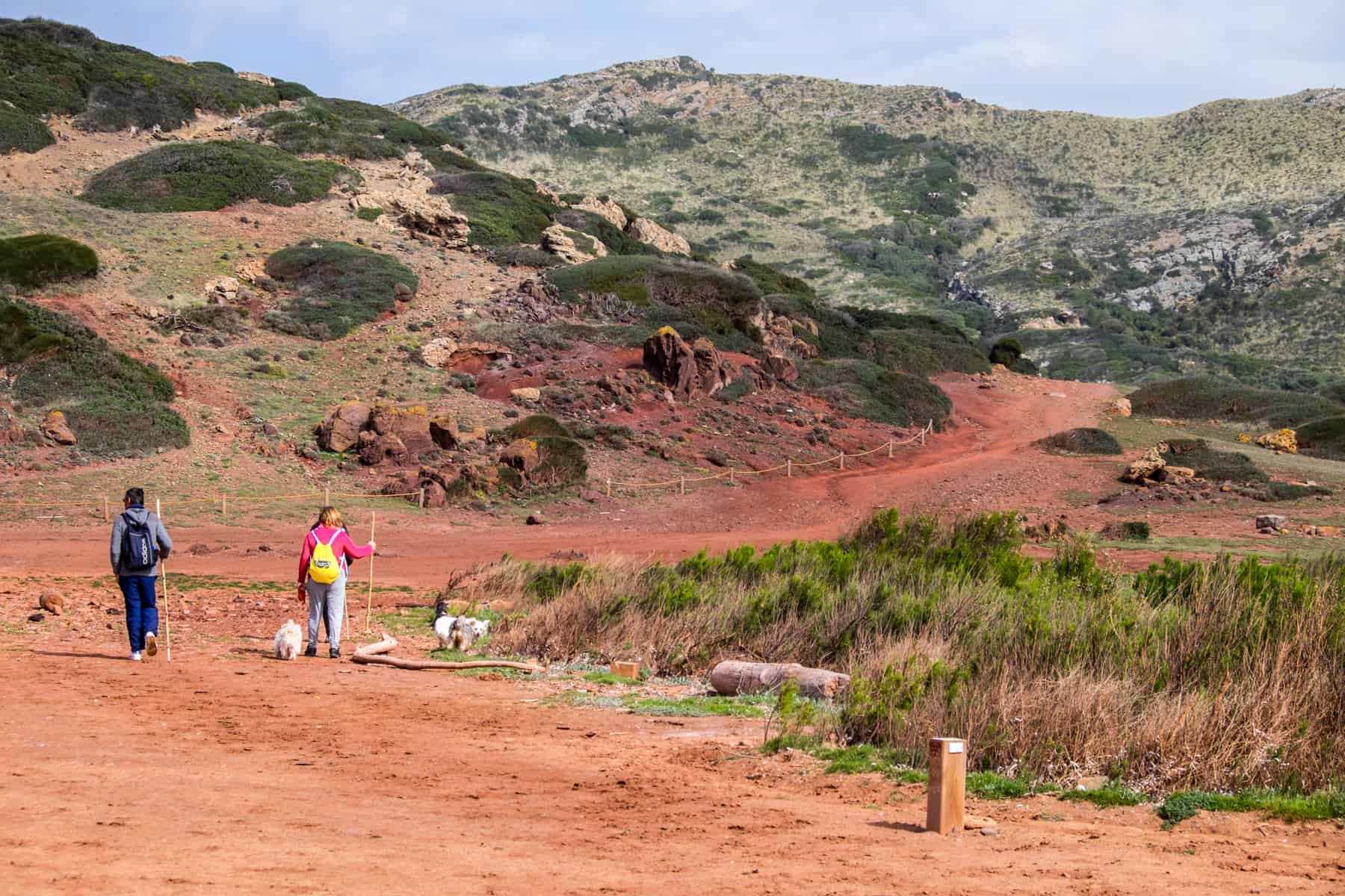 Two walkers with sticks and a dog walk on a sandy orange path towards a small hill and green hills - part of the Cami de Cavalls trail in Menorca. 