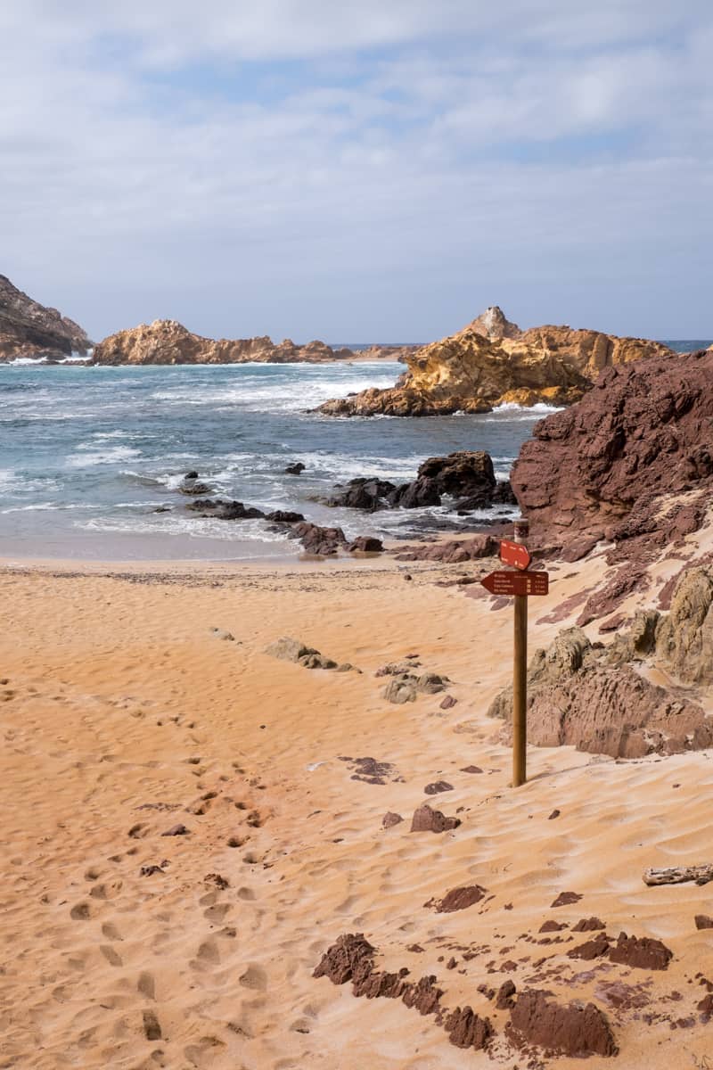 A red walking trail signpost on a sandy, rocky beach pointing the way of the Cami de Cavalls trail in Menorca, Spain