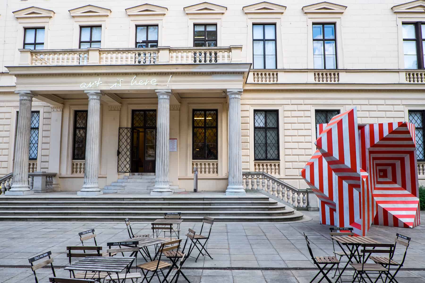 A red and white striped sculpture outside a beige building with white columns with the words 'art is here!' written in a neon light. 