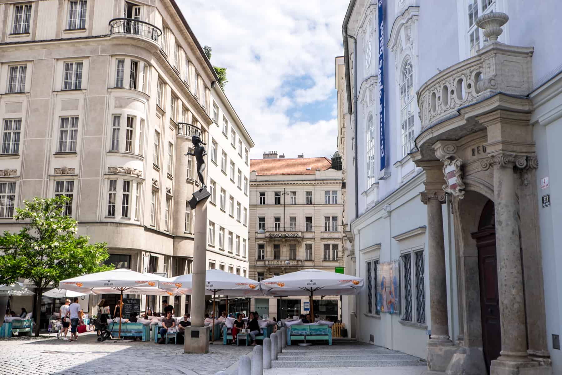 The the statue of Mozart in Brno on a tall column in front of alfreso diners, beige and blue classic style buildings. 