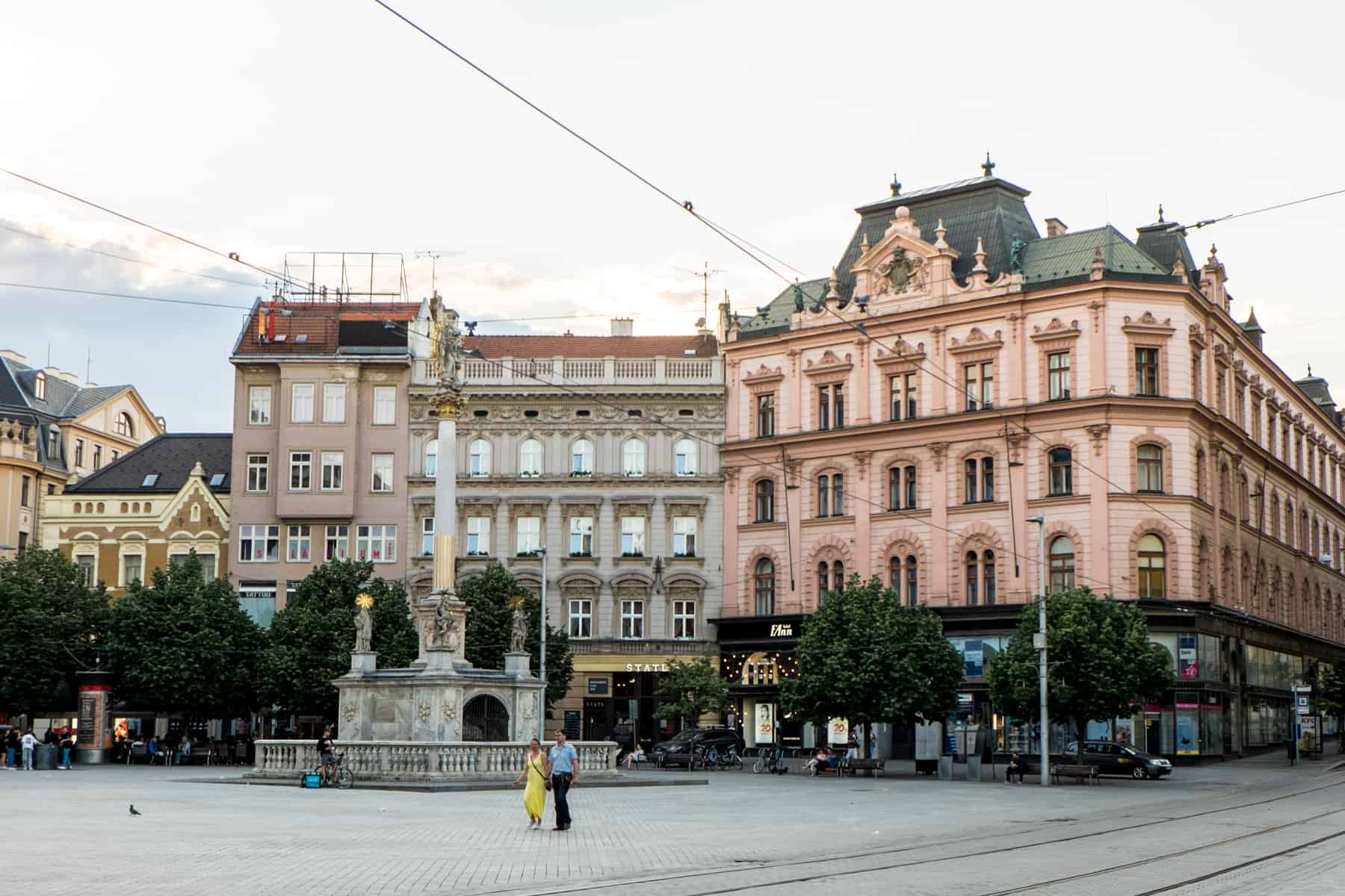 A woman in a yellow dress and a man in a blue shirt walk past a columned fountain in Brno's Freedom Square, in front a large classic looking buildings in varying shades of pink. 