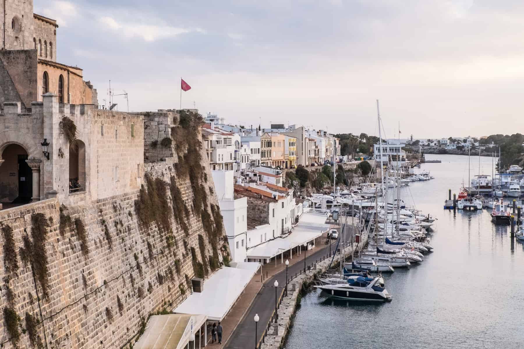 Old golden walls at the port of Ciutadella in Menorca, lined with white buildings and a line of sailing boats on the water. 