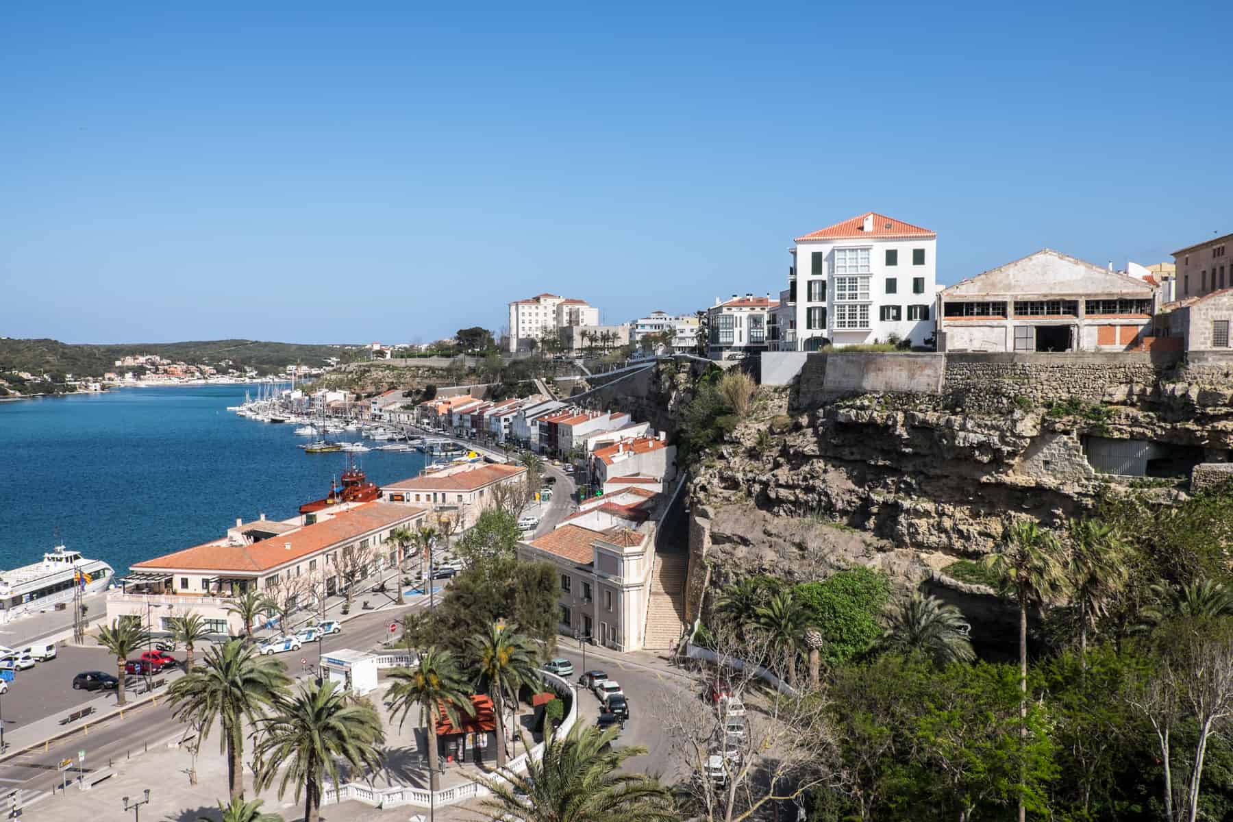 An elevated view over the Port of Mahón in Menorca Islands, looking over old city walls, rows of white houses with range roofs and the rich blue sea bay. 