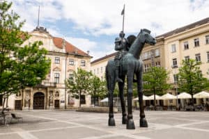 A statue of a man on a horse with giraffe type legs, in a square of golden buildings in Brno, Czech Republic.