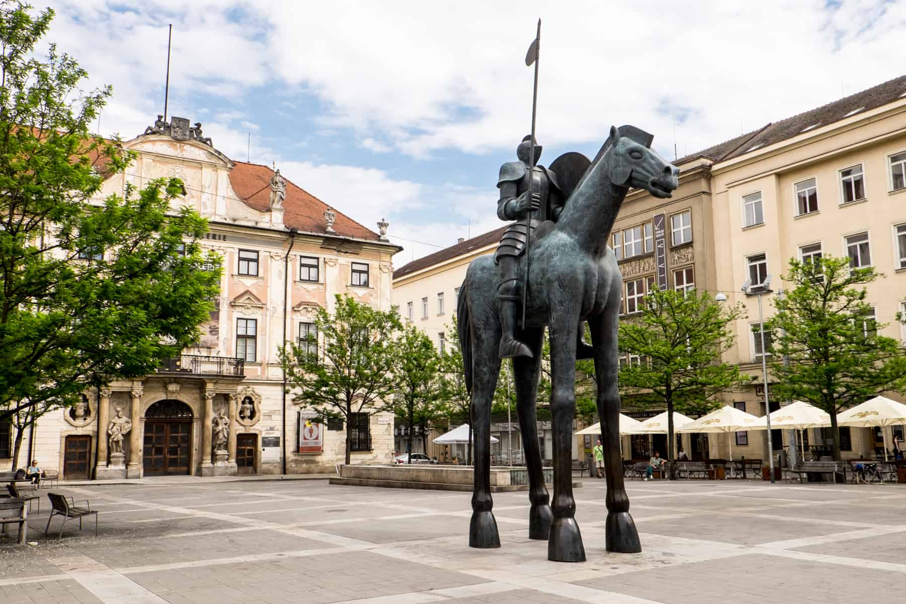 brno places to visit A statue of a man on a horse with giraffe type legs, in a square of golden buildings in Brno, Czech Republic.