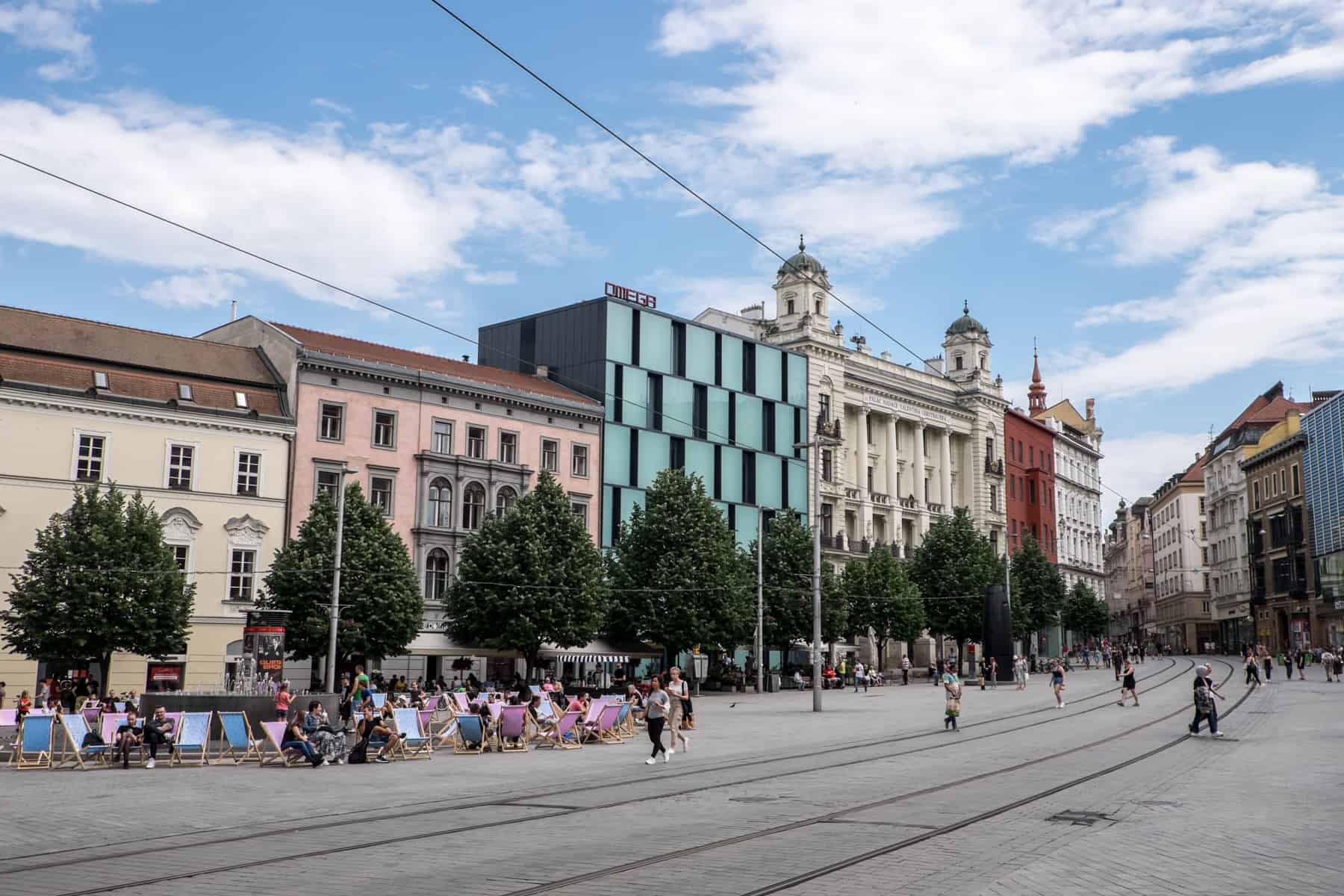 In Brno city, a green functionalist building with the words OMEGA on the roof, wedged between more classic beige, pink and white buildings. People lounge in deck chairs in front. 