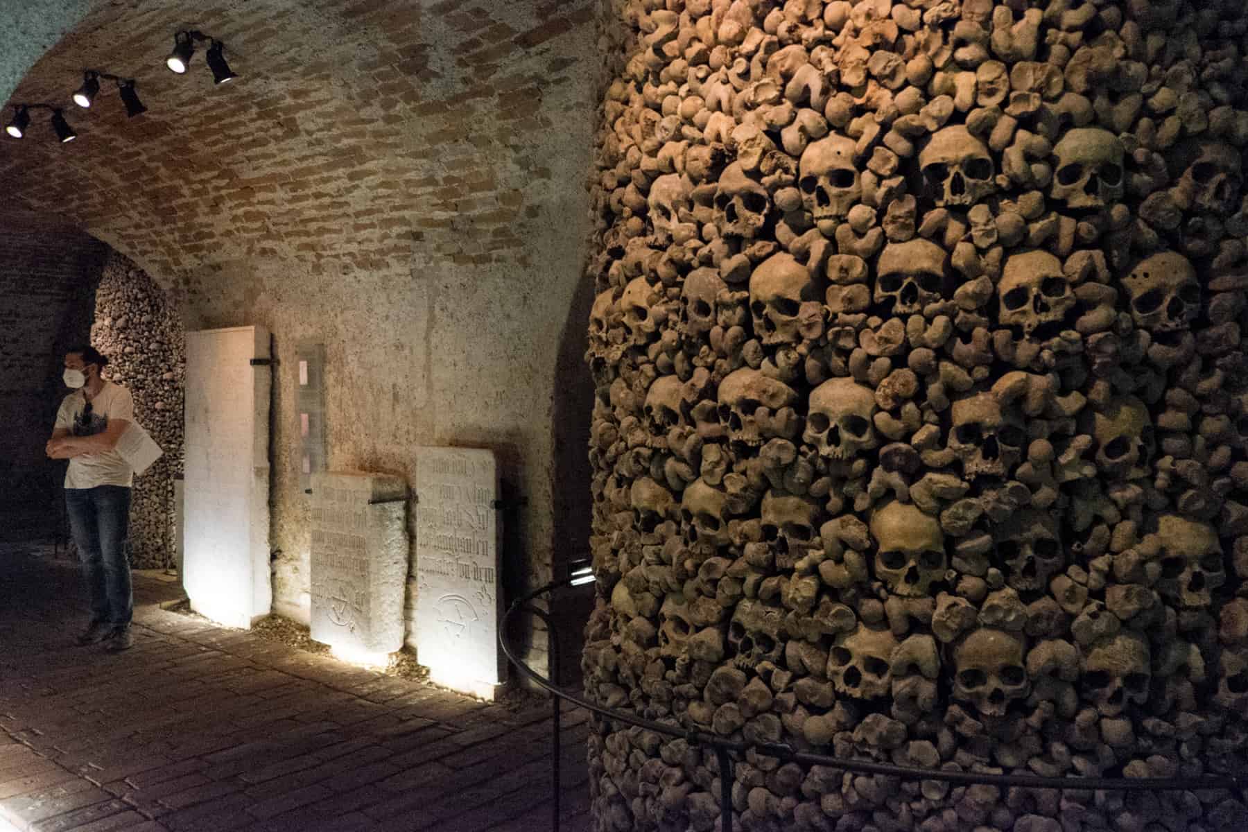 A man stands in from of white tombstones (left) in a room filled with a cylindrical tower of skulls and bones. One of the curious things to do in Brno. 