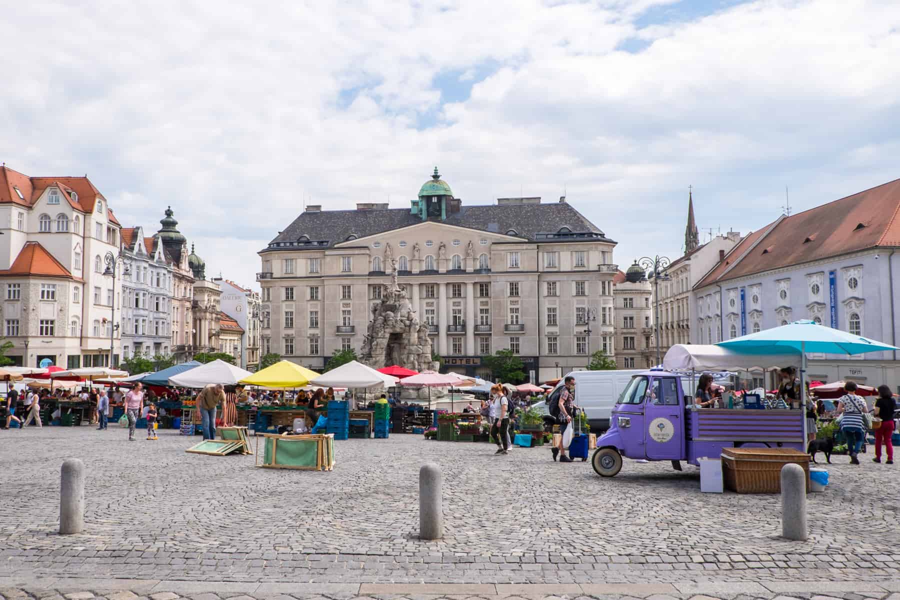 A purple ice cream truck and stalls with umbrellas in rainbow colours fill the Vegetable Market square in Brno surrounded by old buildings.
