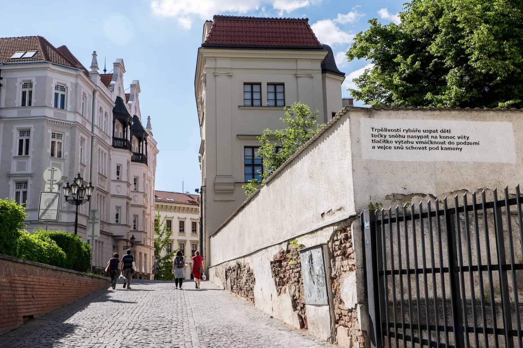 Four people walk up an old stone street in Brno, lined either side with classic, pastel coloured buildings and a crumbling wall.